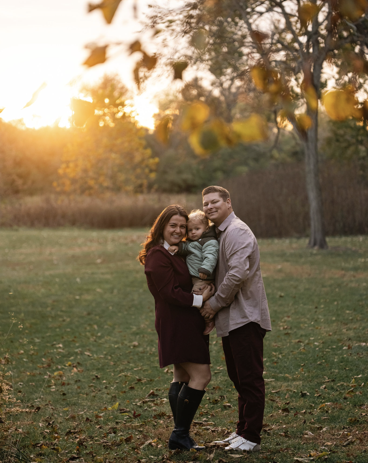 A family of three standing outdoors in a park during sunset, with autumn leaves on the ground and trees in the background. The woman is holding a young child, and a man is holding the child's hand. All are smiling at the camera.