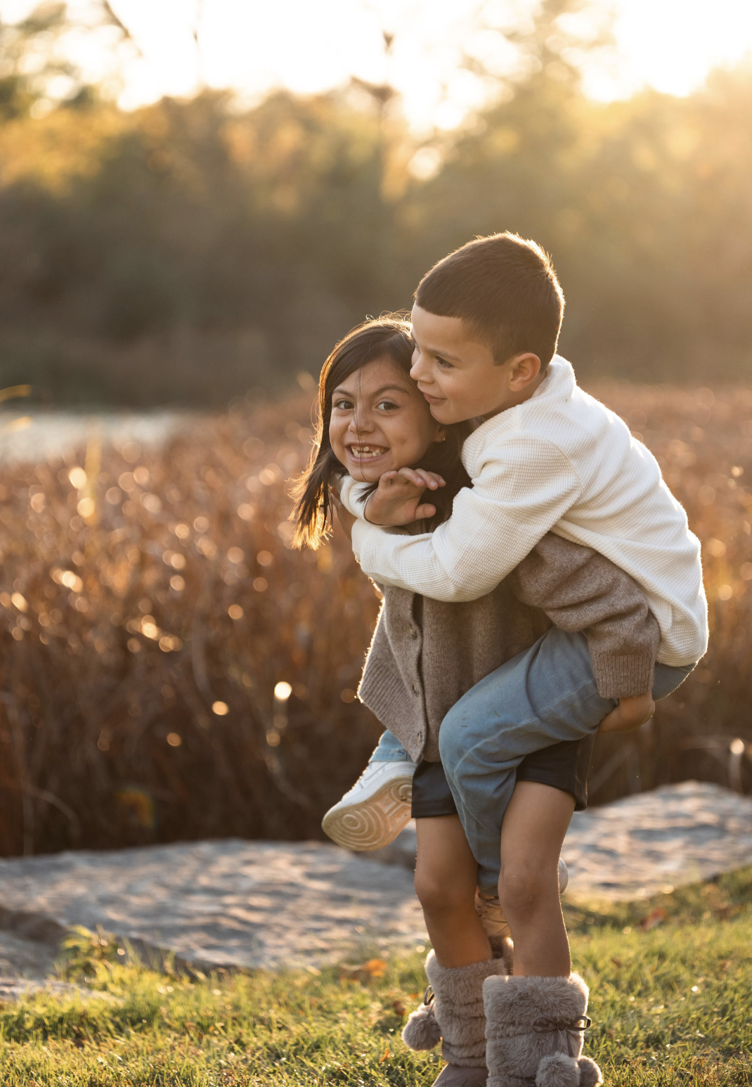 A boy giving a girl a piggyback ride outdoors during sunset, with warm autumn foliage in the background.