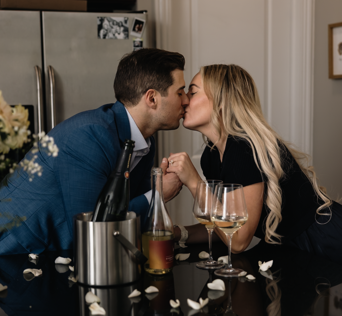 A man and a woman kissing at a dinner table with wine glasses and a champagne bottle, in a cozy indoor setting.