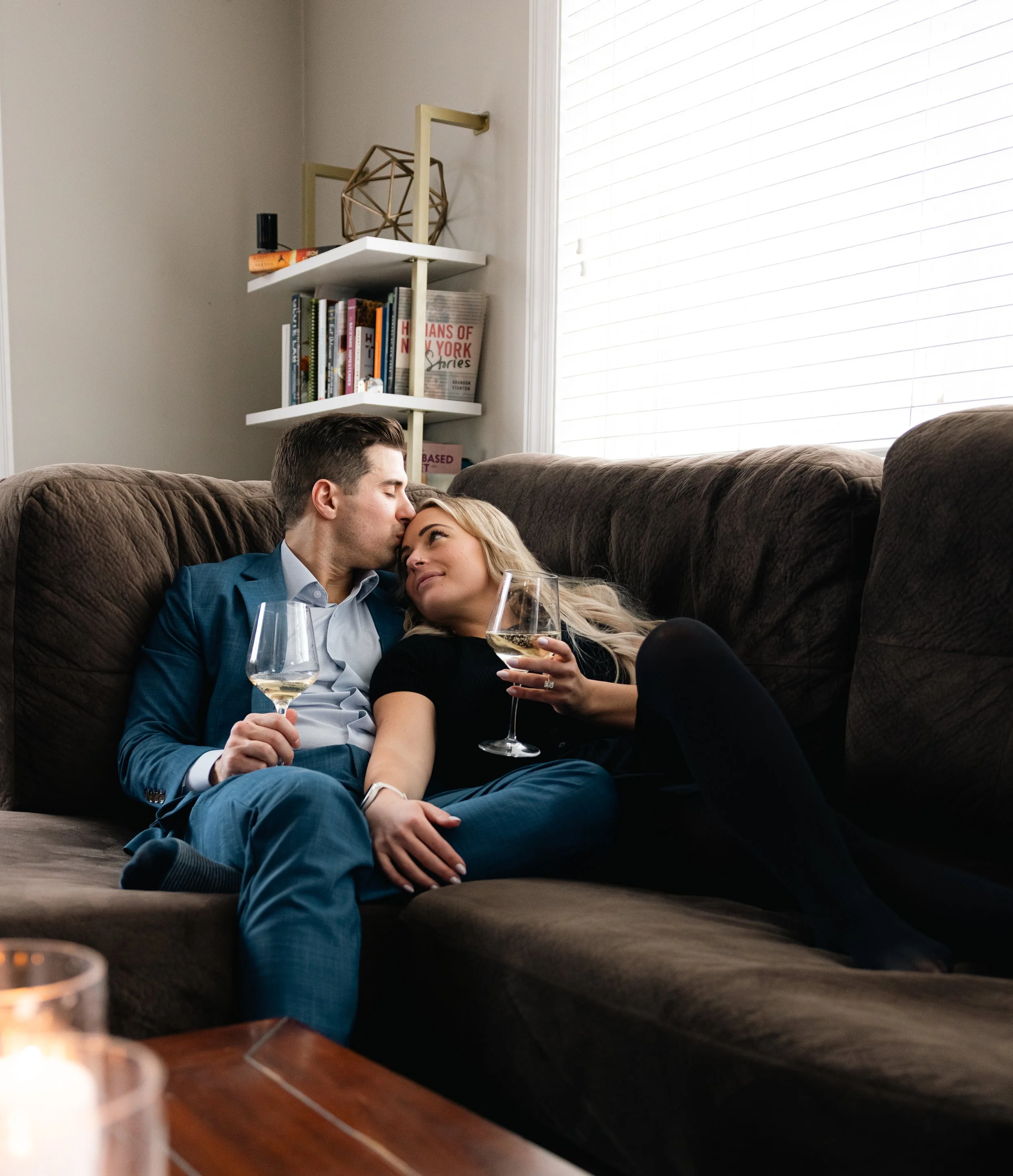 A man and woman sitting closely on a brown couch, holding glasses of white wine, with the man gently kissing the woman's forehead, in a cozy living room with white blinds covering a window and a bookshelf decorated with books and decorative objects b