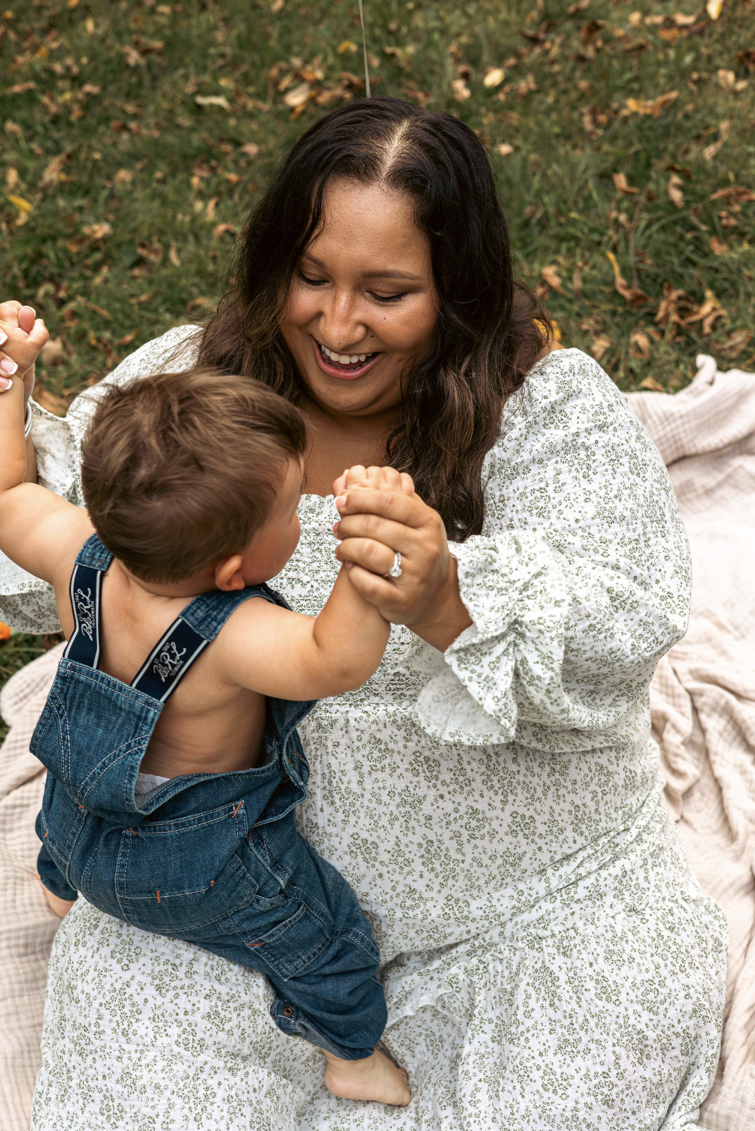 A woman with long dark hair and a white dress with small green floral pattern, smiling, playing wrestling with a small boy with short light brown hair, wearing denim overalls, outdoors on a blanket with fallen leaves on grass.