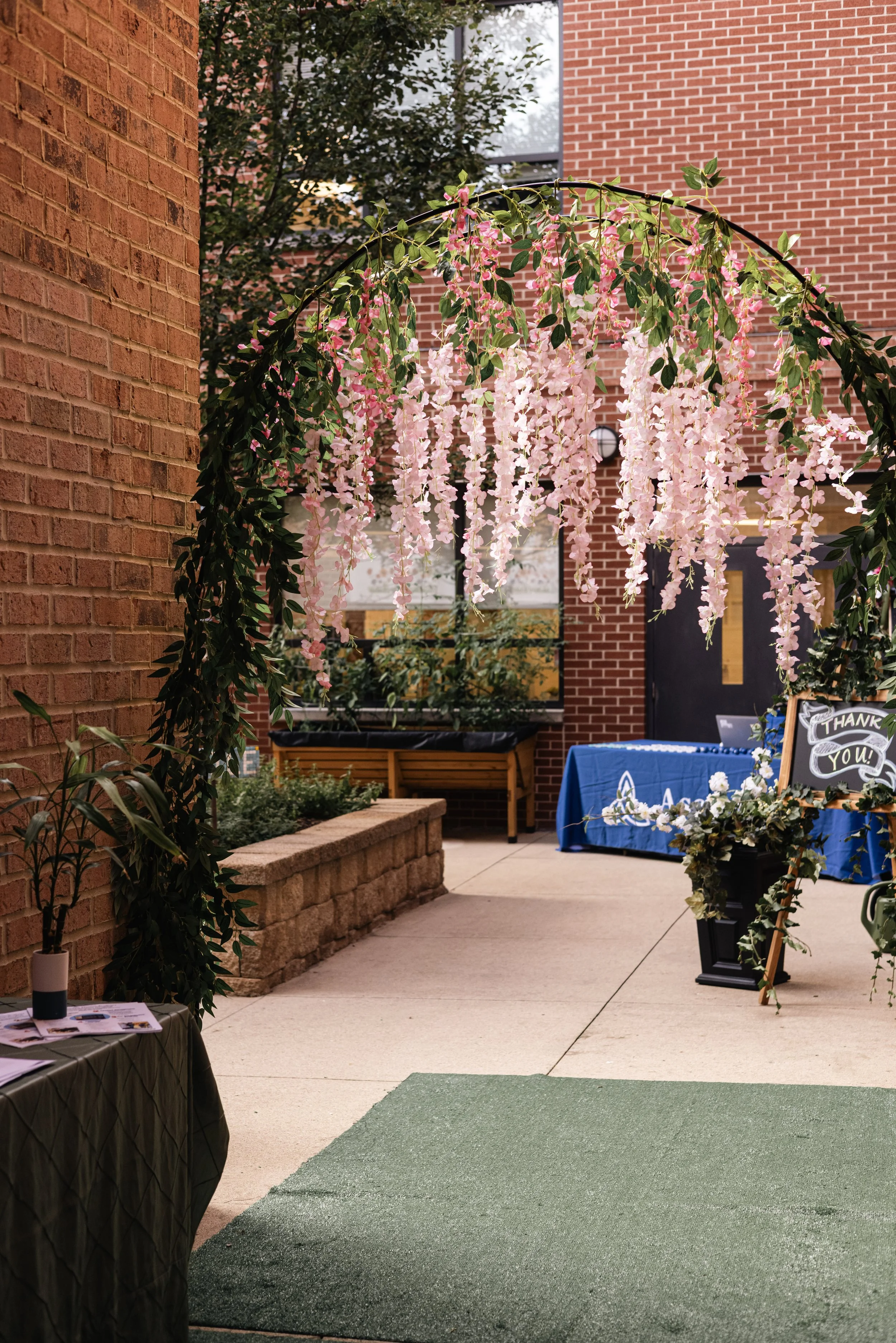 Outdoor area with brick walls, a hanging pink and white flower arrangement, a blue tablecloth with a thank you sign, and a green mat on concrete ground.