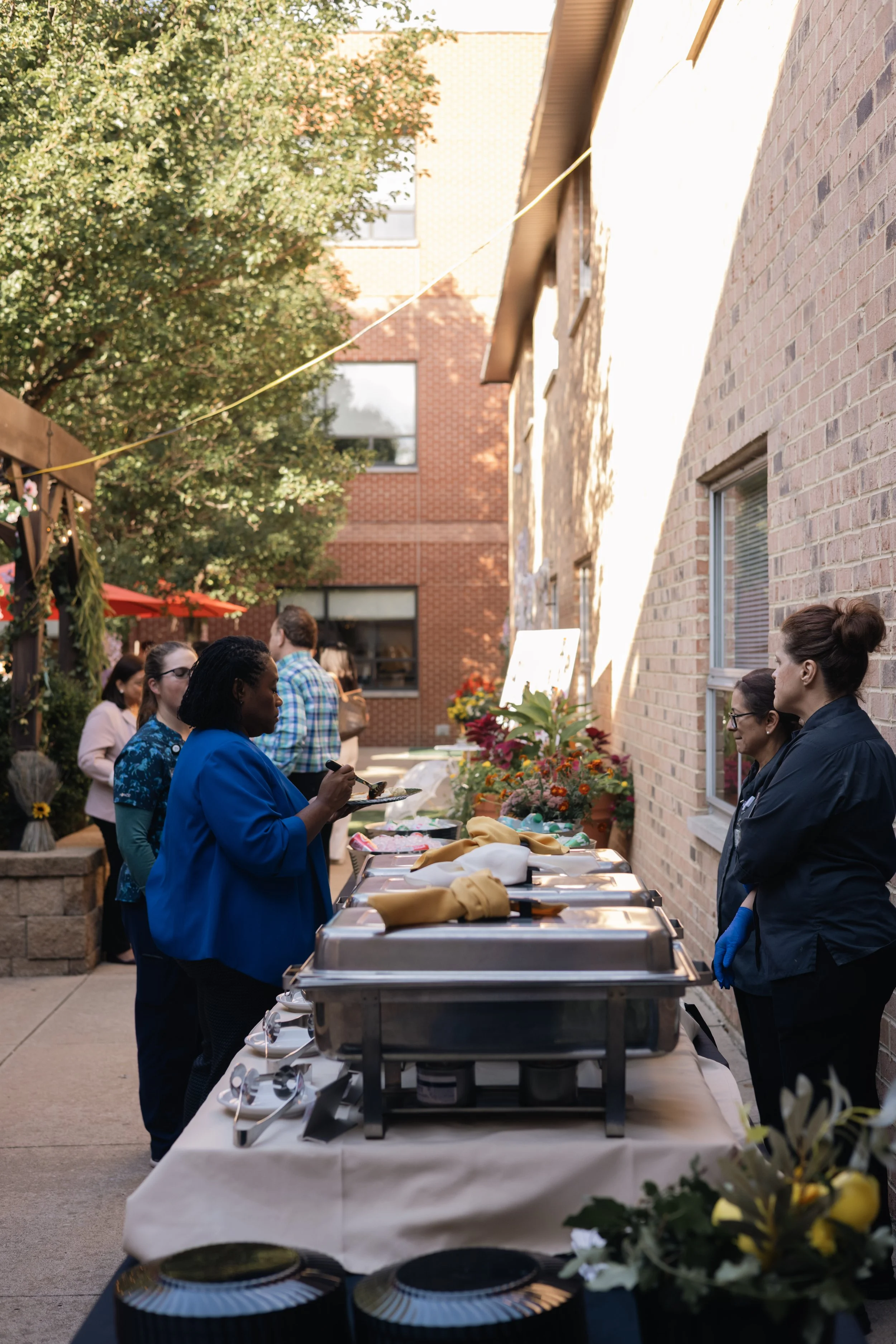People at an outdoor event near a brick building, standing around a buffet table with various dishes and decorations, some wearing gloves.
