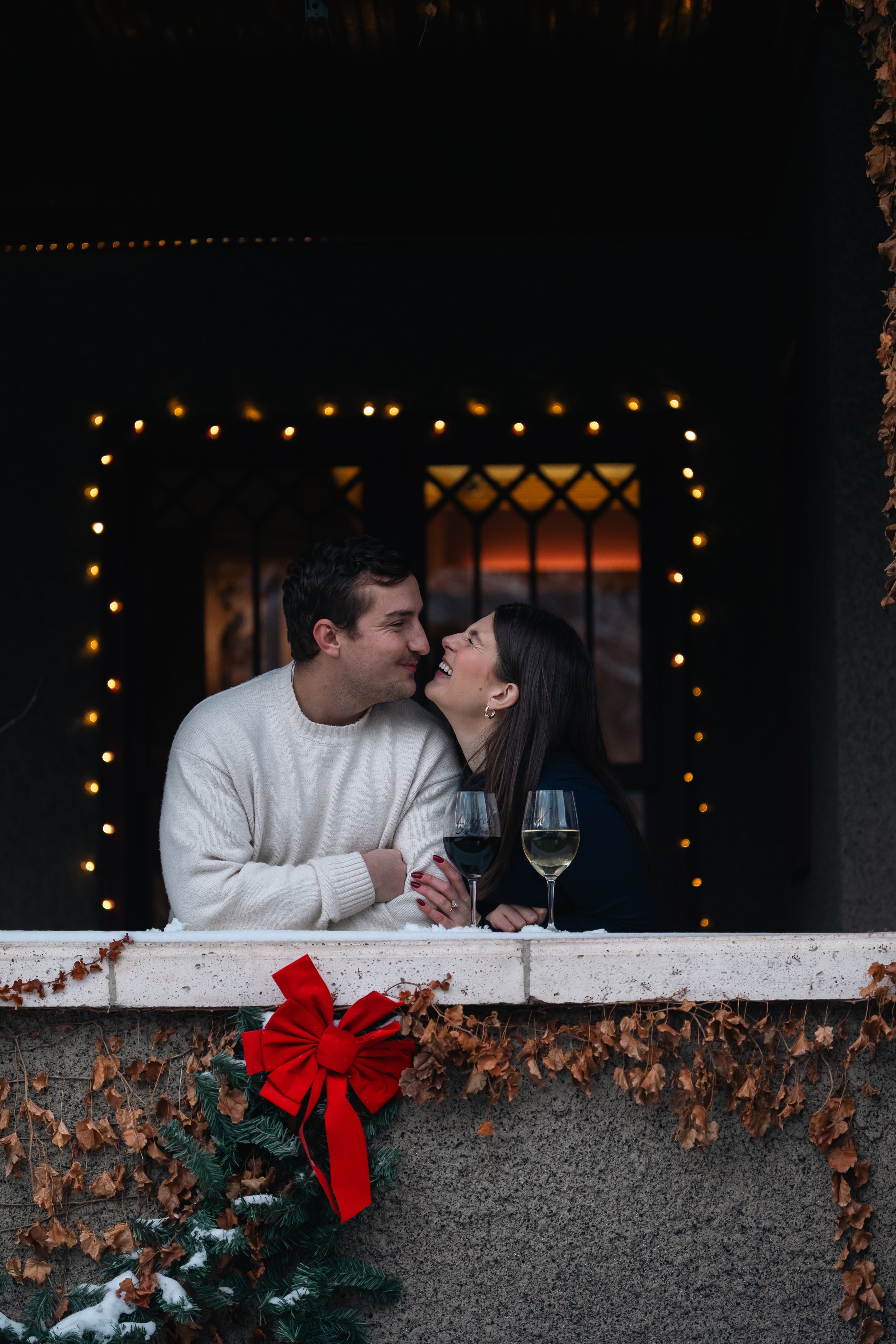A man and woman sharing a kiss and smiling, holding glasses of white wine at a decorated Christmas holiday gathering, with Christmas lights and a wreath with a red bow in the background.