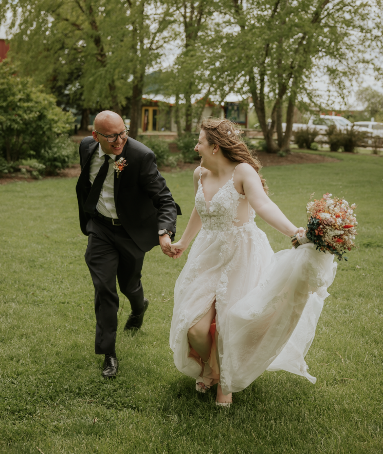 A bride and groom holding hands and running across a grassy area outdoors during a wedding celebration, with trees and a park in the background.