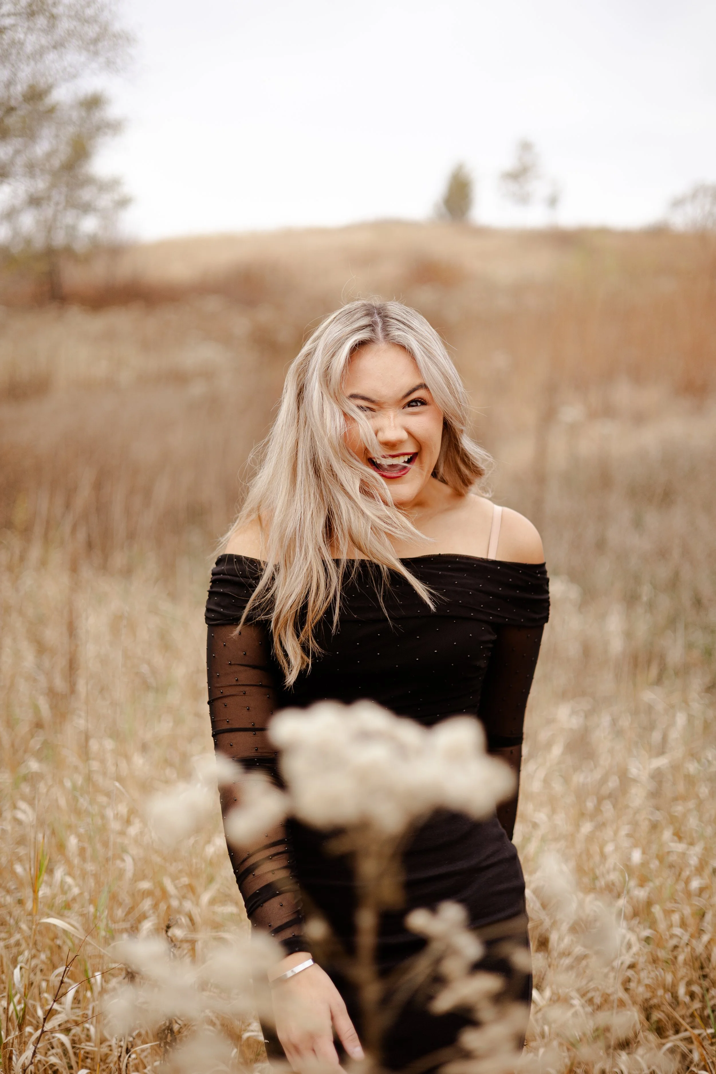 A young woman with blonde hair making a playful face in a field of tall, beige grass, wearing a black off-shoulder dress with sheer sleeves.