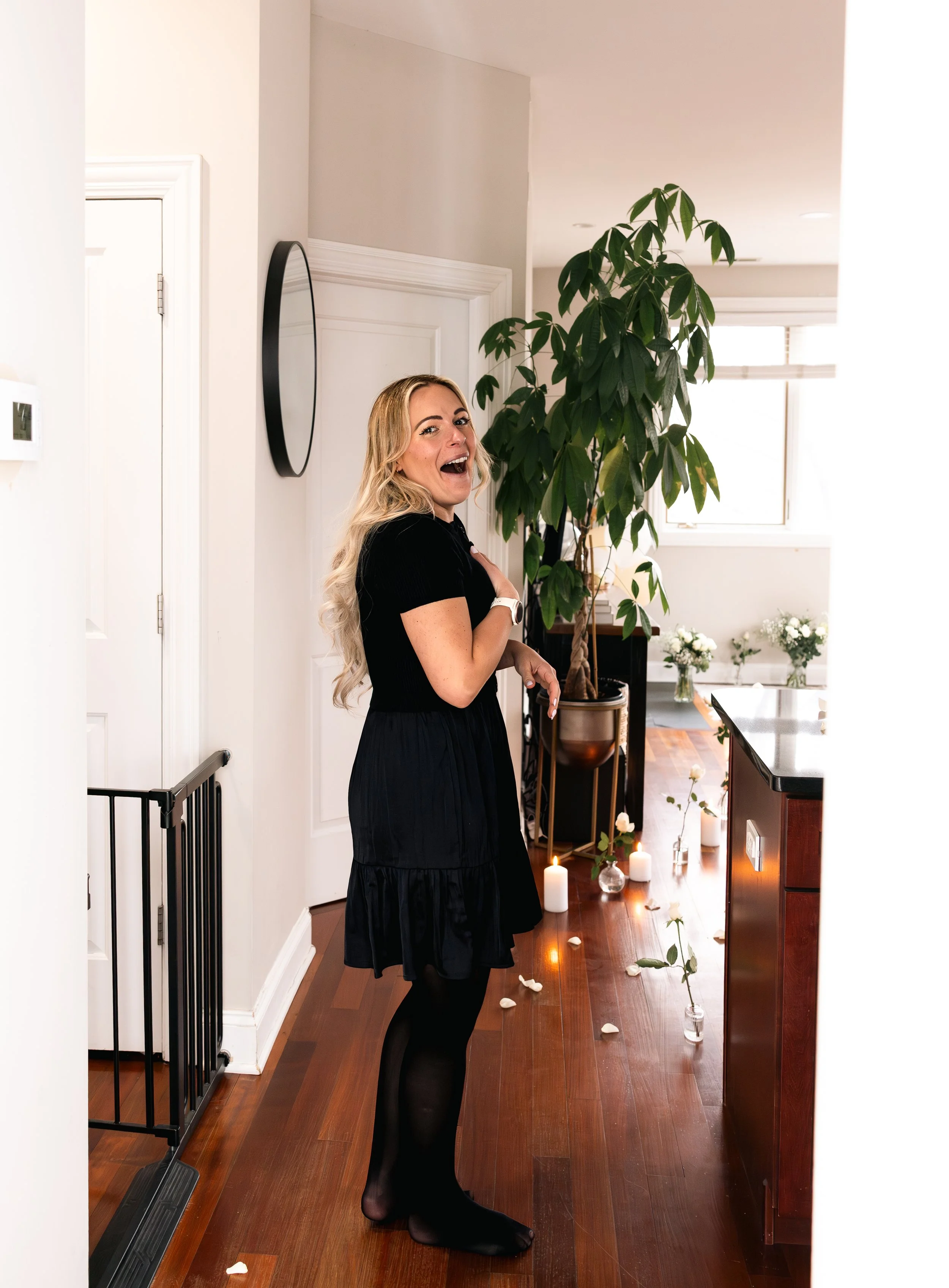 A smiling blonde woman with long hair, wearing a black dress and black tights, standing inside a warmly lit room with candles and flowers, next to a tall green plant.