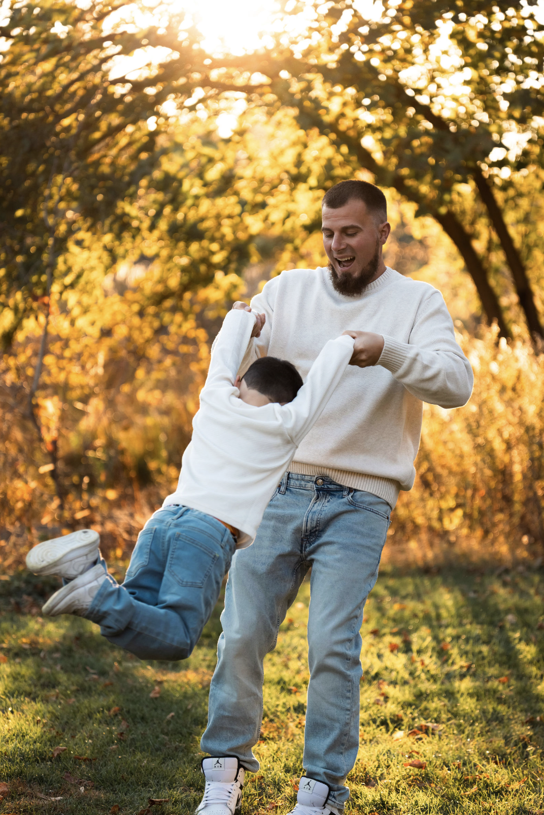 A man and a boy playing and having fun outdoors in a park during autumn, with golden sunlight filtering through trees in the background.