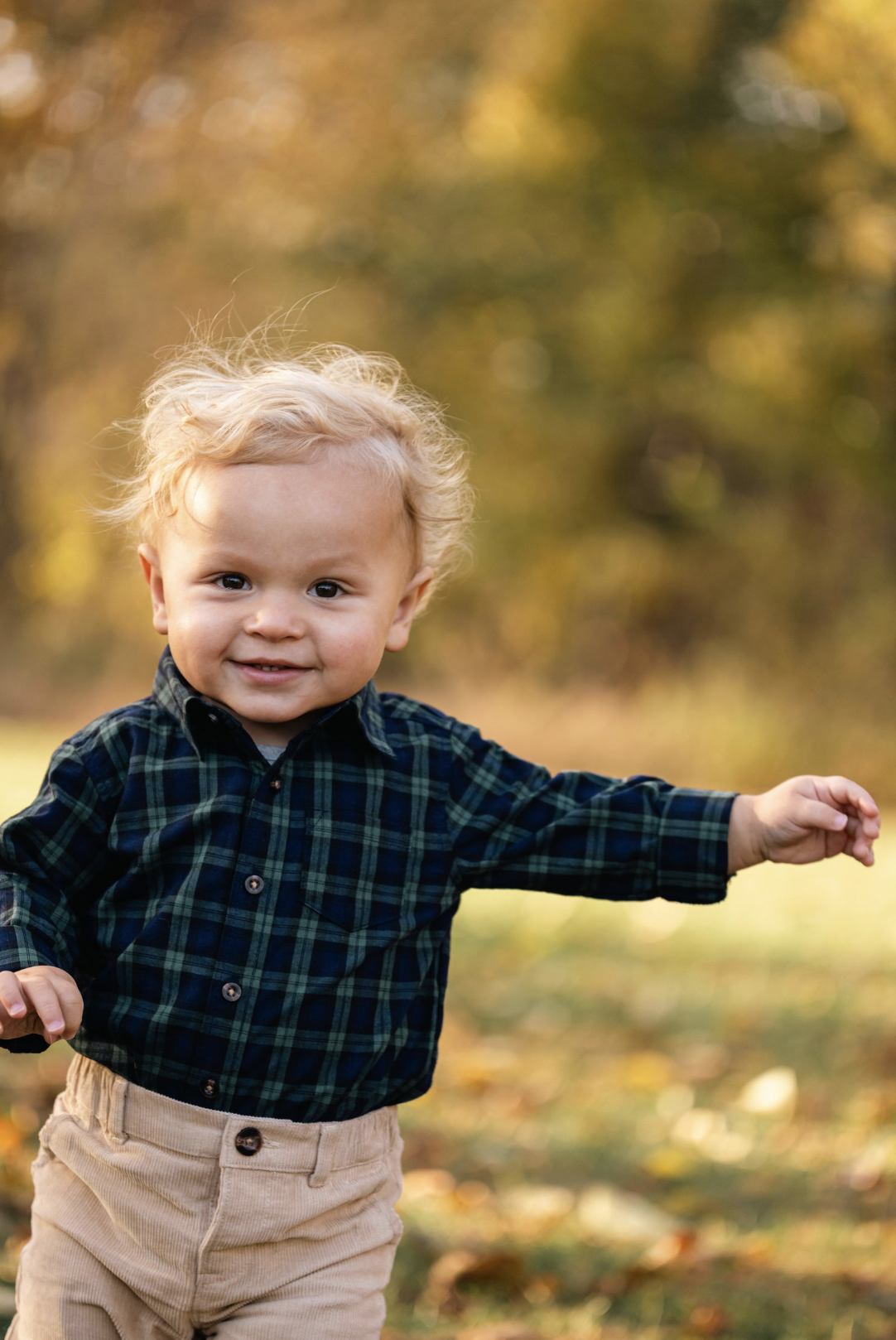 A young boy with curly blonde hair and a plaid shirt standing outdoors in a park with fall foliage.