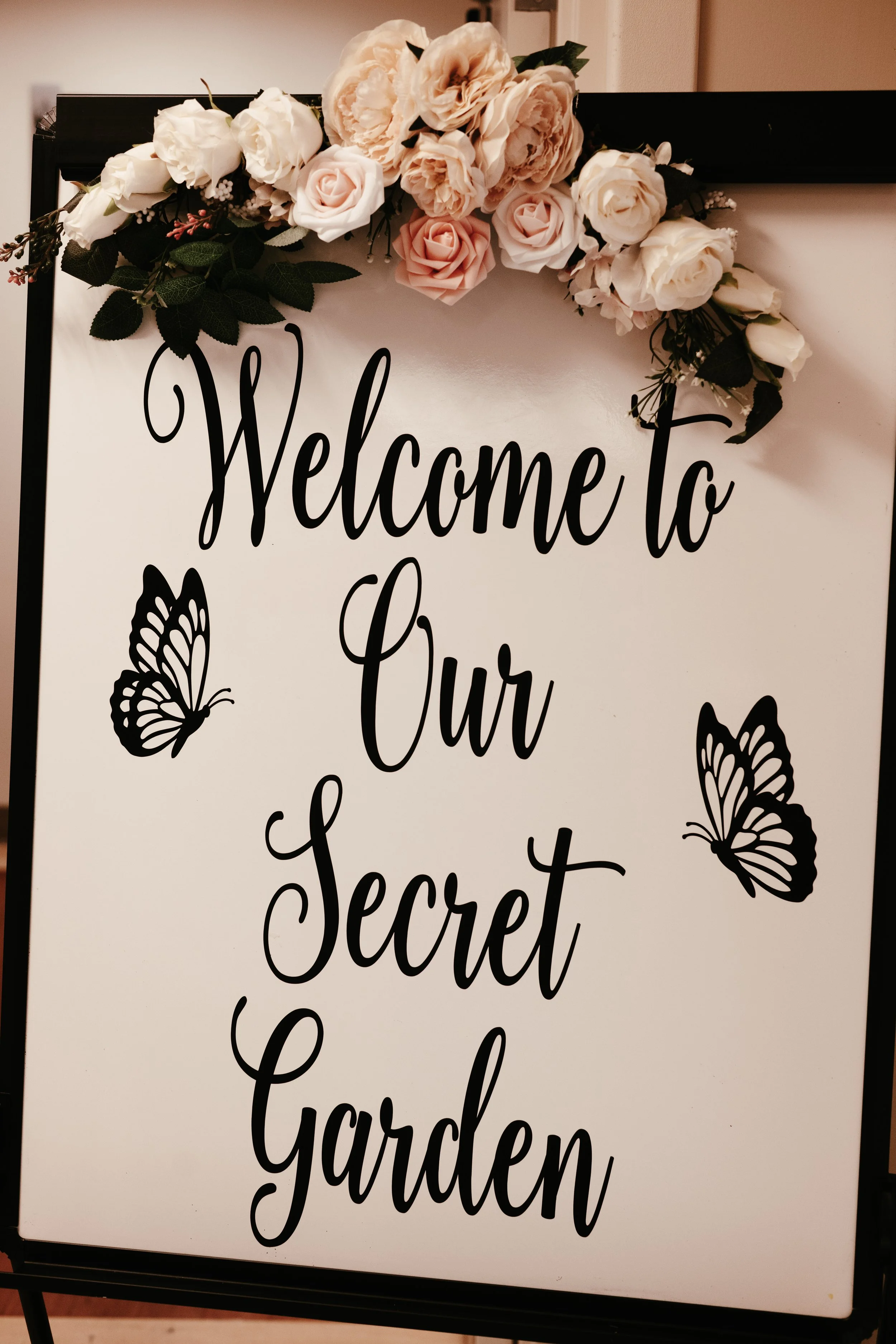 Sign with black floral and butterfly decorations on top, reading 'Welcome to Our Secret Garden' in elegant black script.