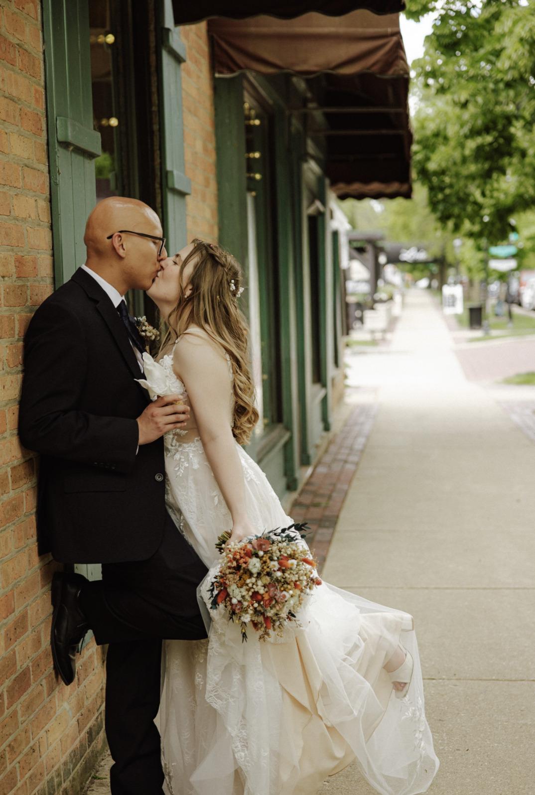 A newlywed couple sharing a kiss on a city sidewalk. The groom is dressed in a black suit and glasses, and the bride is wearing a white wedding dress and holding a colorful bouquet of flowers.