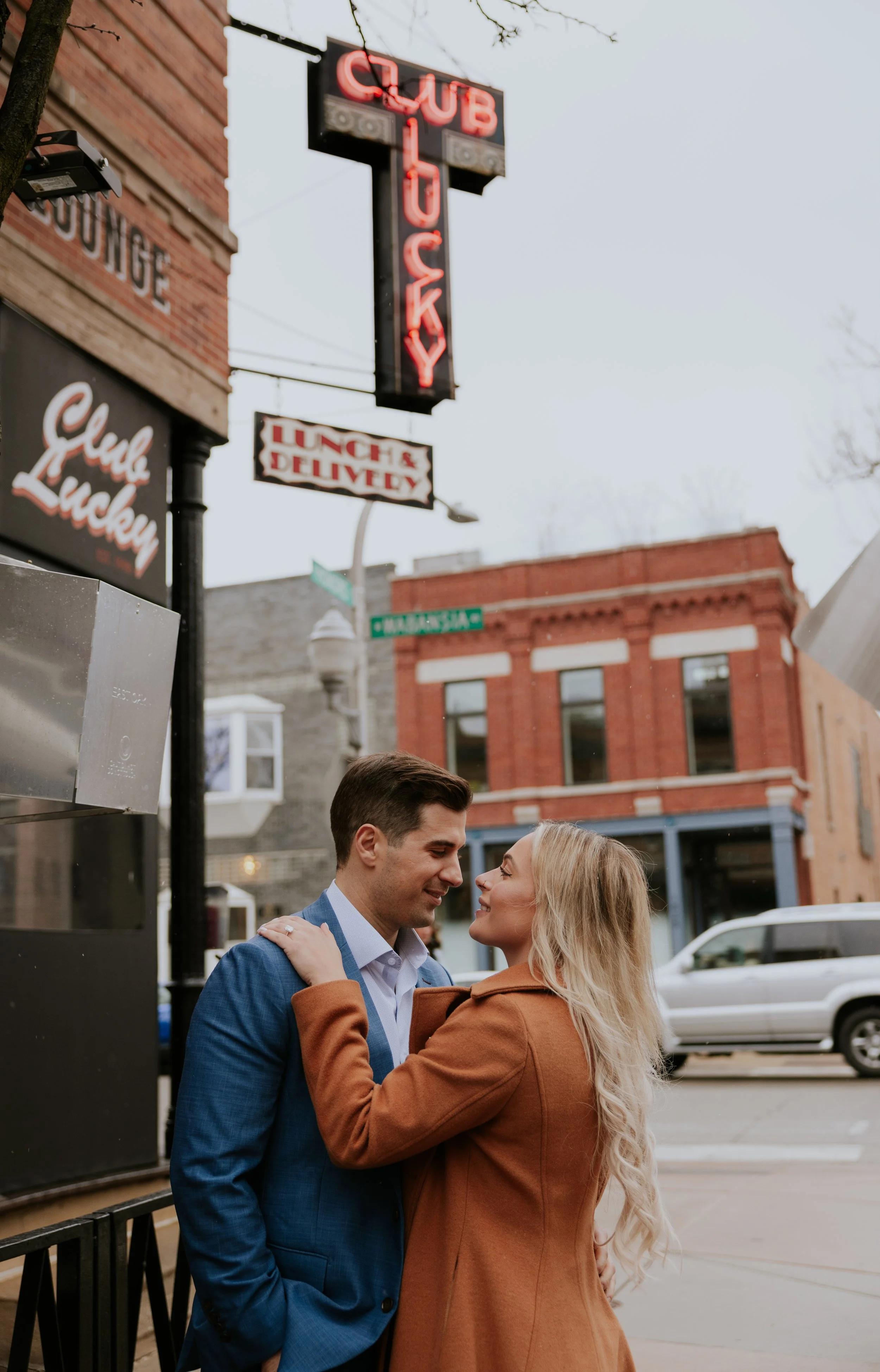 A couple standing close together, smiling and gazing into each other's eyes on a city street with brick buildings and signs that read 'CUB.' one sign says 'LUNCH & DELIVERY' and another says 'Club Lucky.'