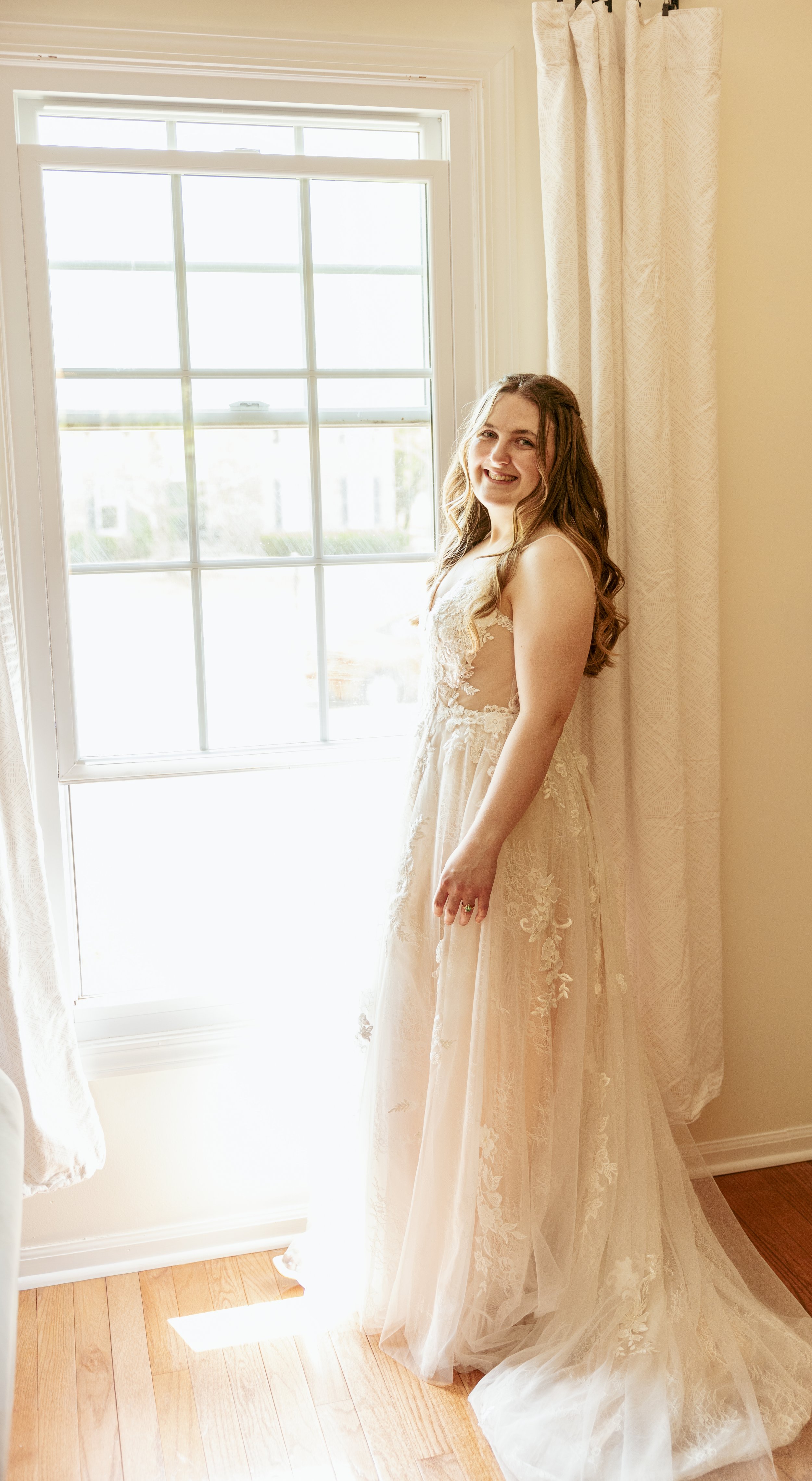 A woman in a wedding dress standing by a window with curtains, smiling at the camera.