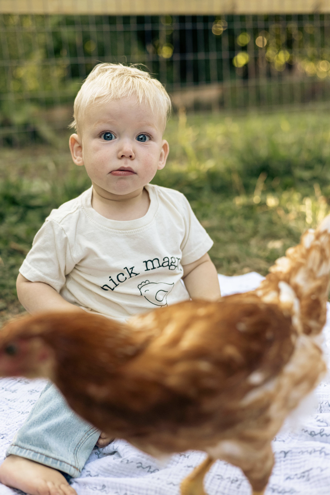 A young blonde-haired boy with blue eyes sitting outdoors on a blanket, wearing a cream t-shirt with a bear and the text 'nick mag' on it. A chicken is in the foreground.