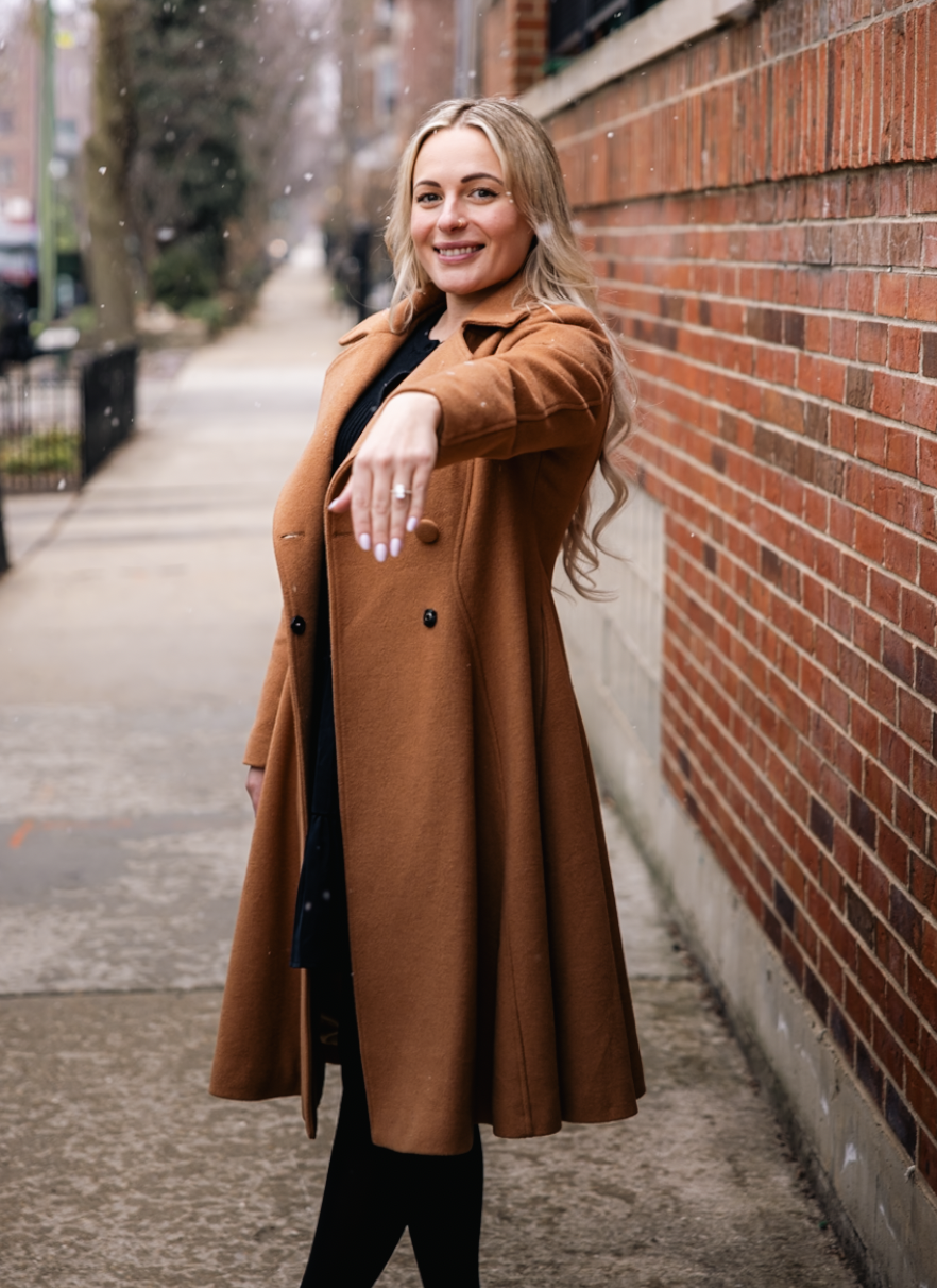 A smiling woman with long blonde hair pointing towards the camera, wearing a tan coat and black clothing, standing on a sidewalk next to a red brick wall in an urban setting.