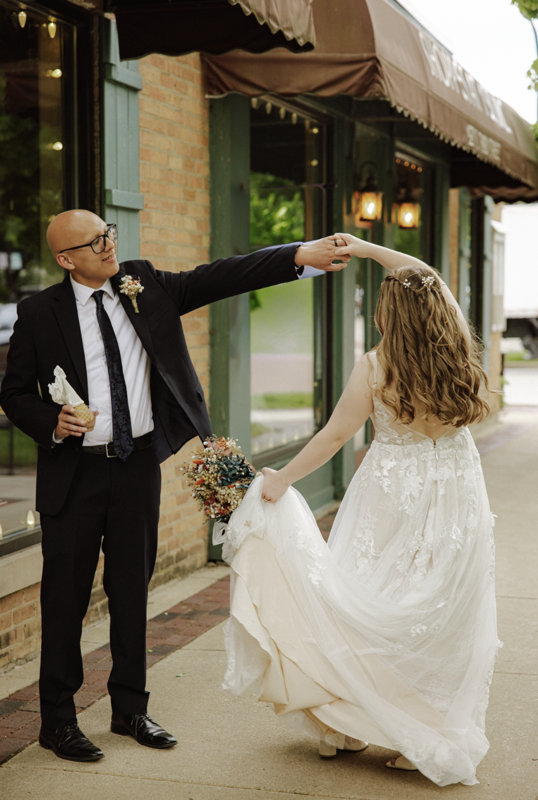 A groom in a black suit and glasses is holding a cone of ice cream and smiling while holding hands with a bride in a white lace wedding dress, who is lifting her dress to show her bouquet as they enjoy a moment together outside a restaurant or cafe.