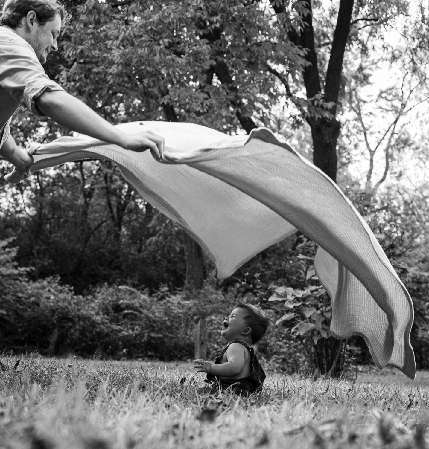 A man is playfully lifting a blanket or sheet over a young child sitting on the grass in a park. Both are outdoors with trees and foliage in the background. The child is laughing and looking up at the man.