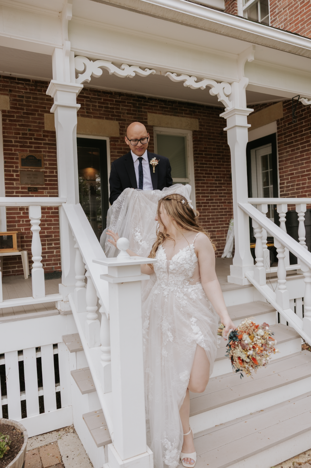 A bride in a lace wedding dress holding a bouquet of flowers stands on stairs, with a groom in a suit holding a lace veil above her head, on a white porch with brick walls in the background.