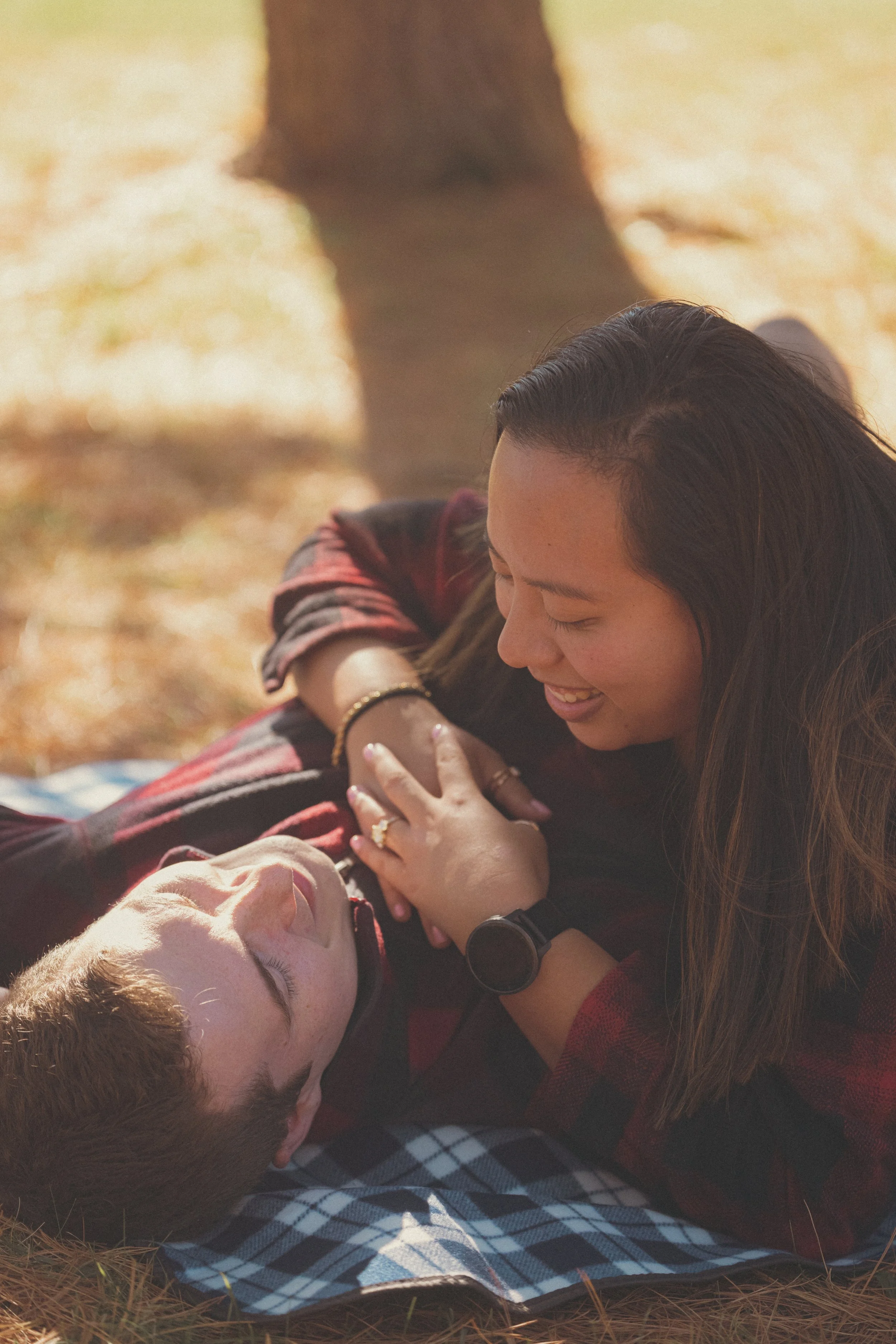 A woman and a man are lying on the ground outdoors, smiling and looking at each other affectionately. The woman is leaning over the man, who is lying with his eyes closed. They appear happy and relaxed, with autumn leaves on the ground.