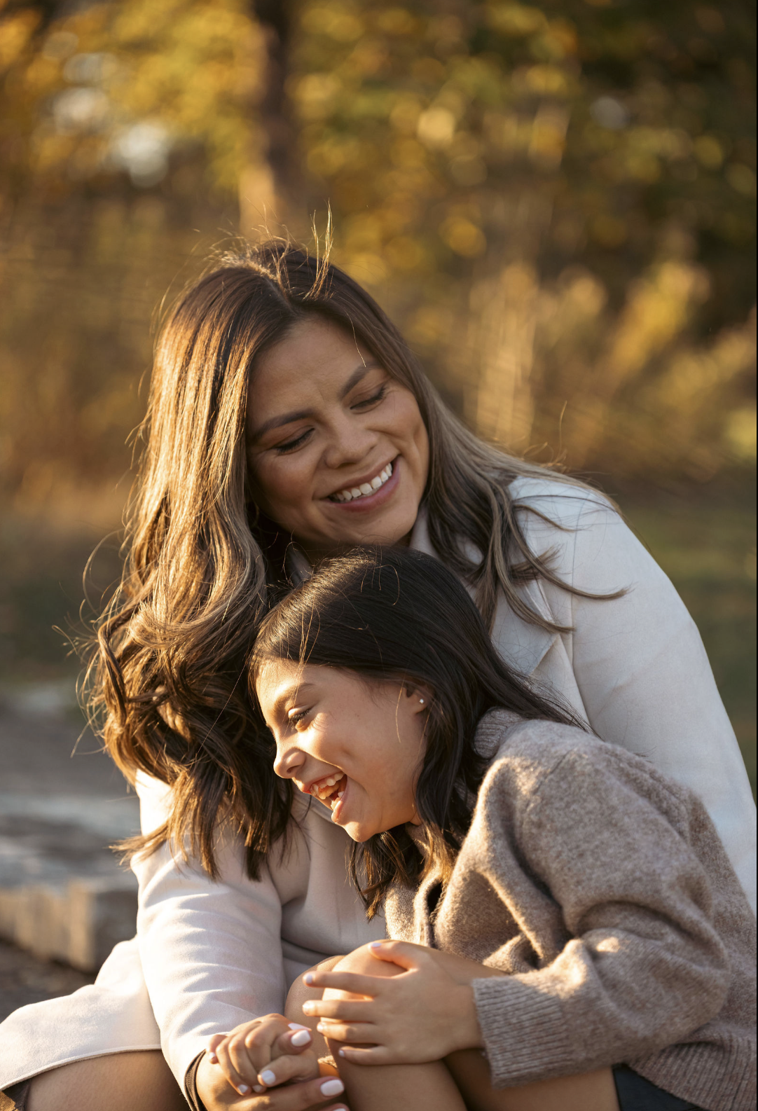 A woman and a girl are laughing and hugging outdoors during golden hour with autumn trees in the background.