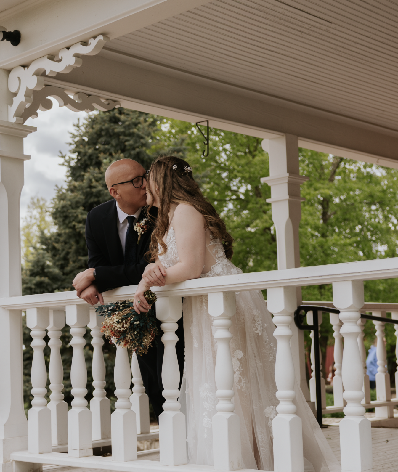 A bride and groom sharing a kiss on a white gazebo, with trees in the background.