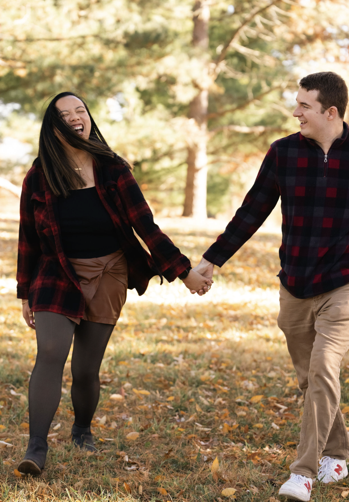 A couple holding hands and walking through a park with fall foliage, smiling and enjoying each other's company.