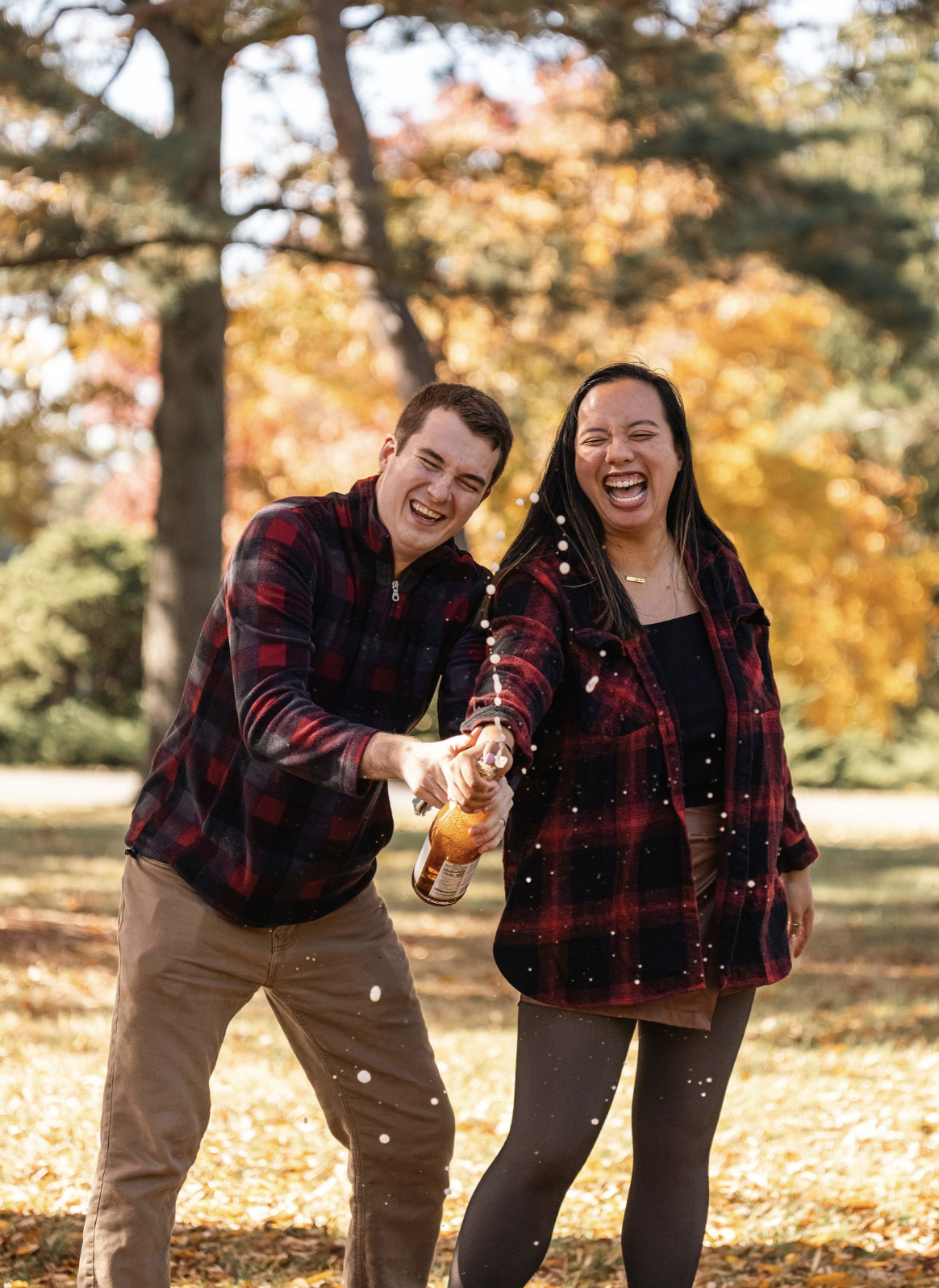 A man and woman celebrating outdoors in a park with autumn foliage, spraying champagne and smiling.