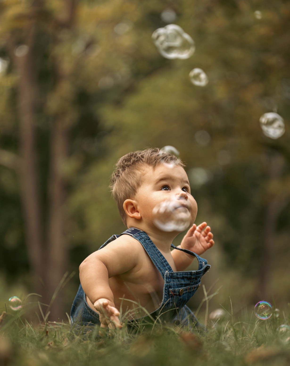 A young boy in denim overalls playing outdoors on a grassy field, surrounded by bubbles, with trees blurred in the background.