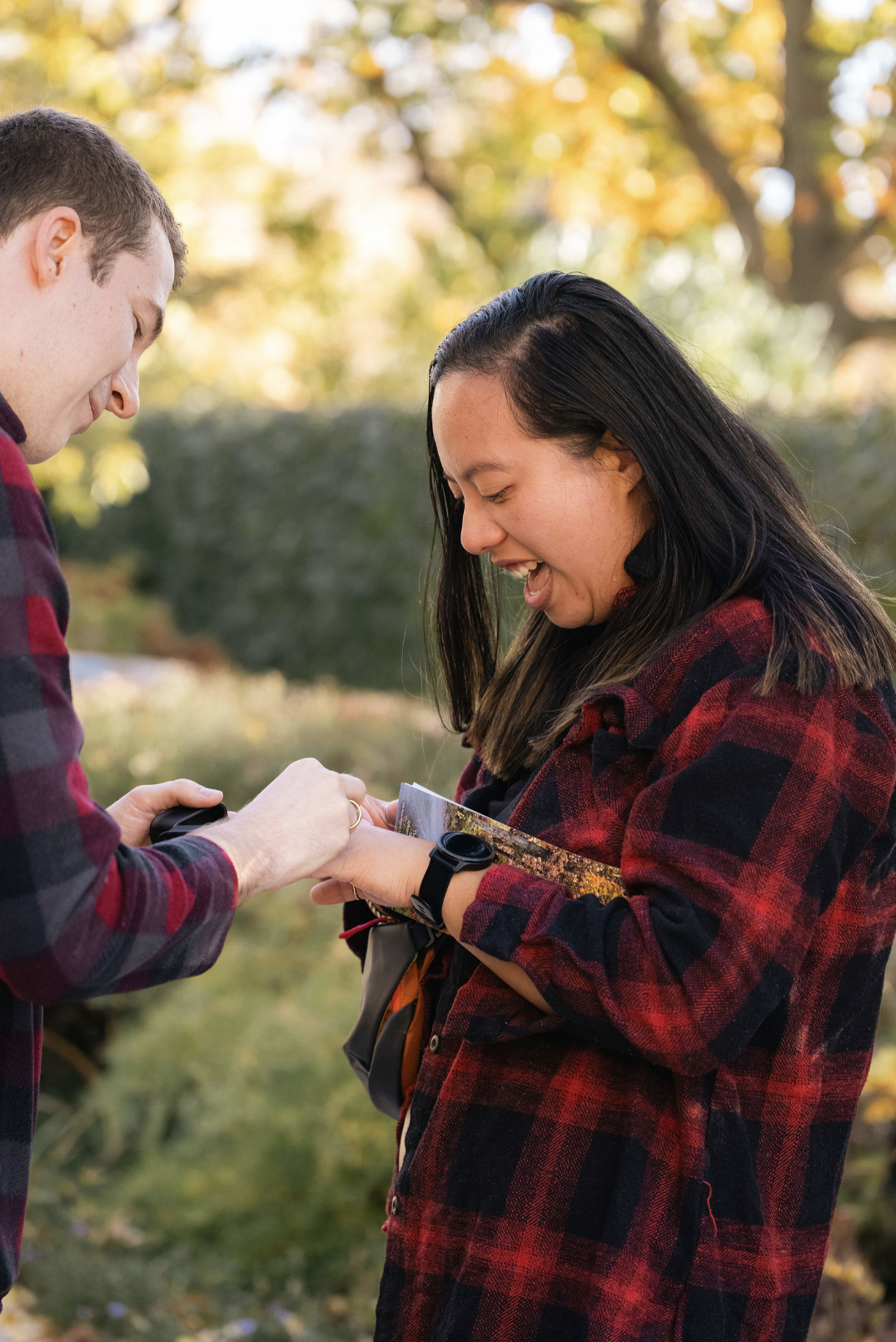 Two people outdoors, one woman and one man, smiling and sharing a joyful moment. The woman is holding a photograph and wearing a black wristwatch, while the man is placing something on her wrist. They are wearing red and black plaid shirts with autum