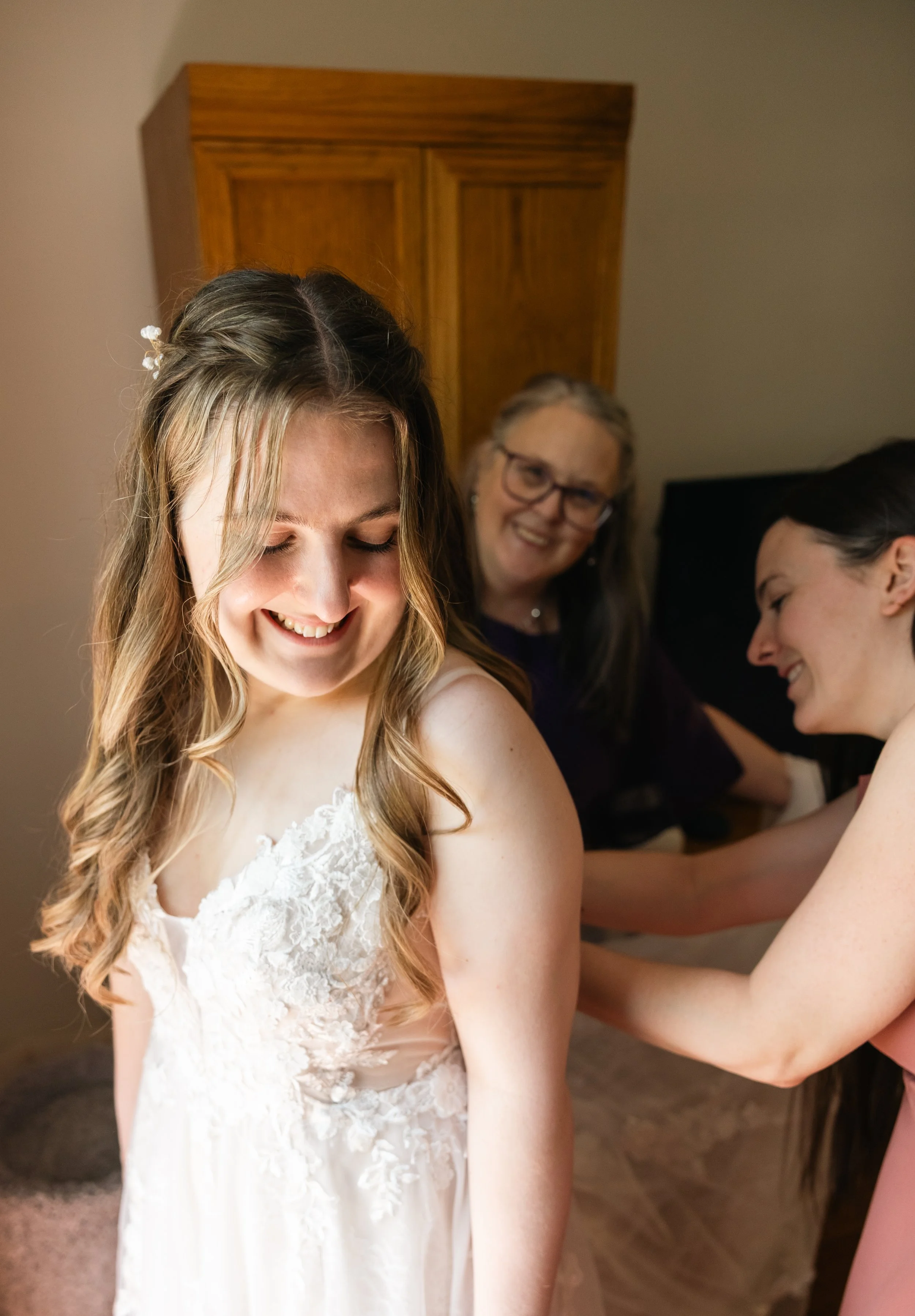 A bride in a white lace wedding dress smiling as she gets ready, with two women helping her in the background.