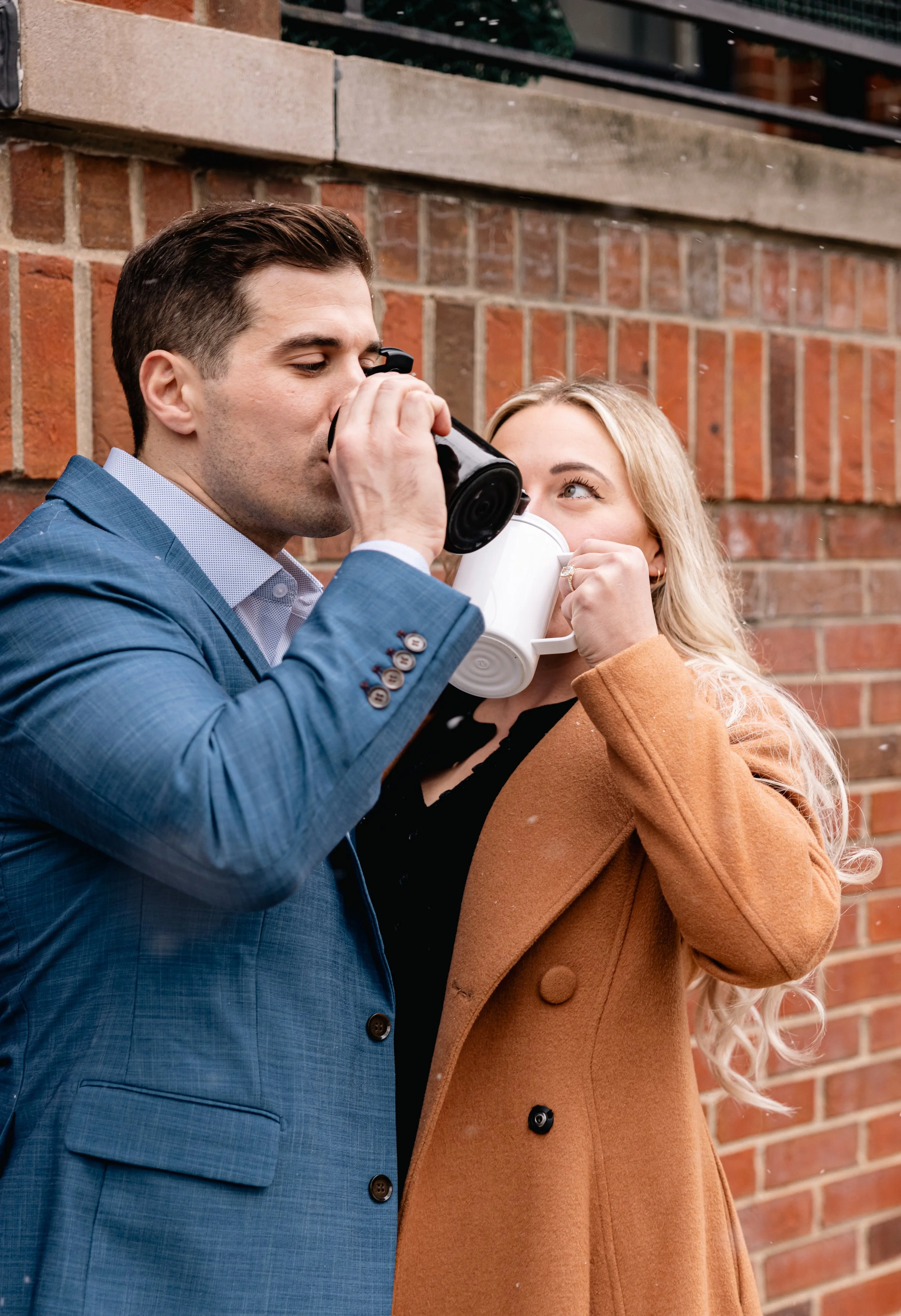 A man in a blue suit drinking from a black travel mug while a woman in a brown coat holding a white mug looks up at him against a brick wall background.