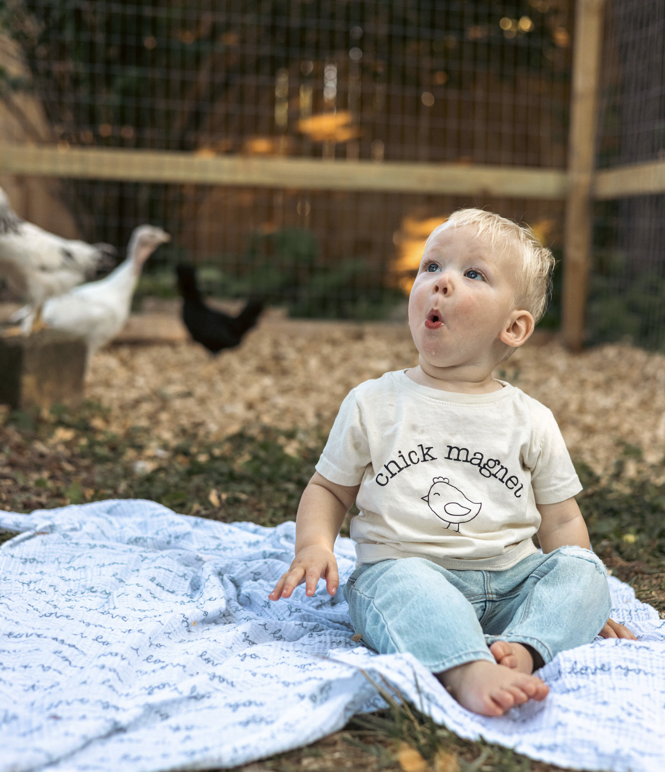 A young toddler sitting on a white blanket outdoors near a chicken coop, with chickens in the background and a blurred wooden fence.