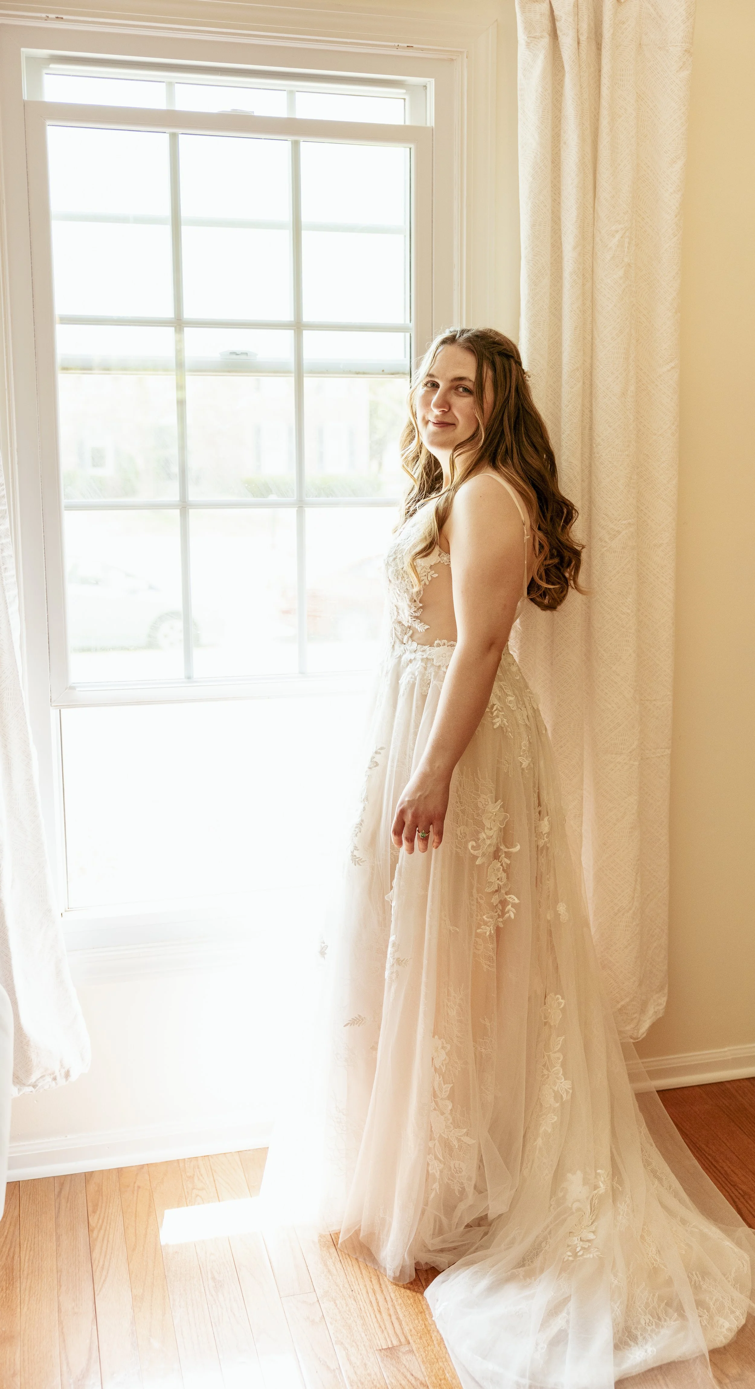 A woman in an elegant, light-colored wedding dress standing next to a window with sunlight streaming in, surrounded by cream curtains and wooden flooring.