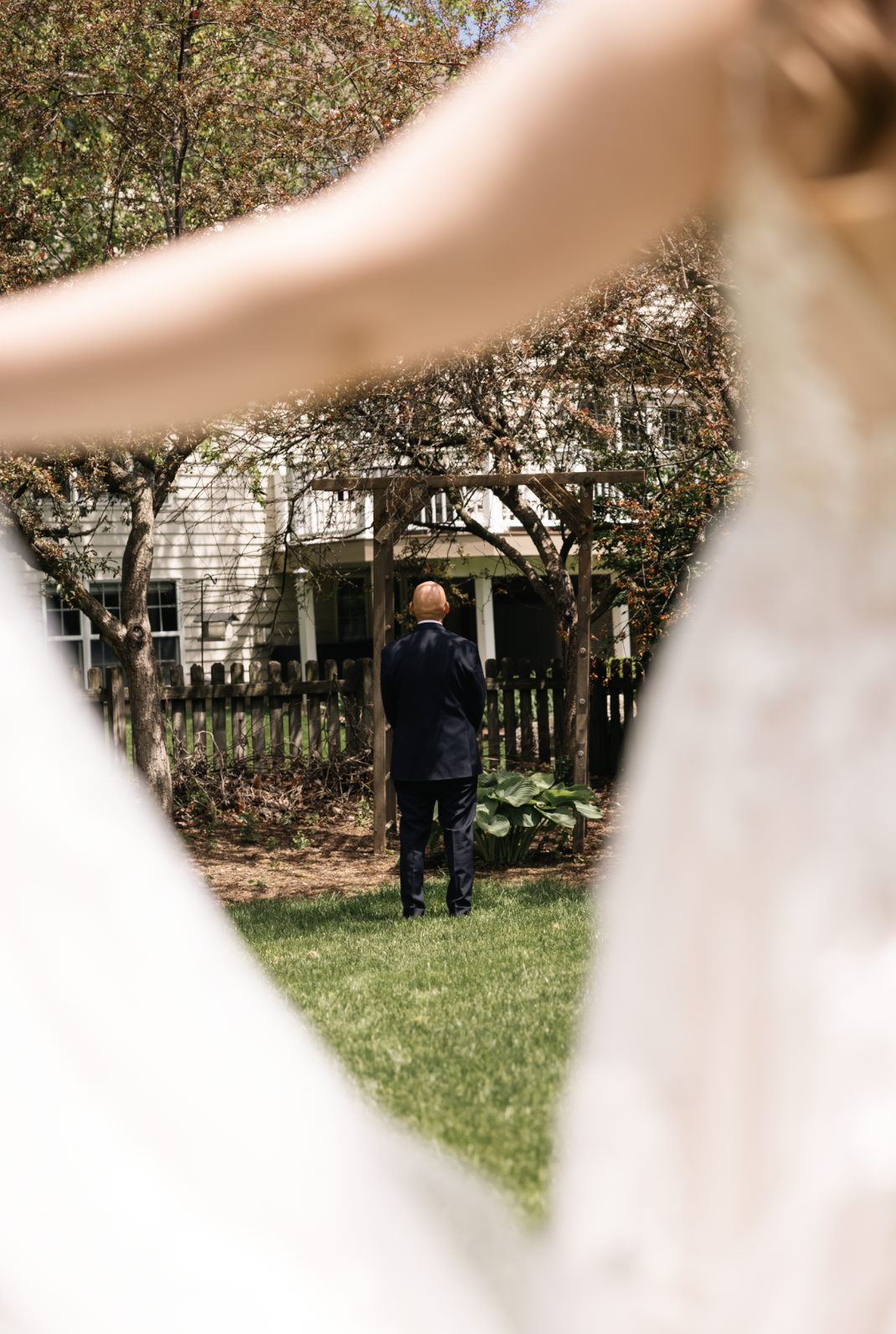 Man in a dark suit standing in a garden, viewed through a white fence with a diamond-shaped opening, with trees and a house in the background.