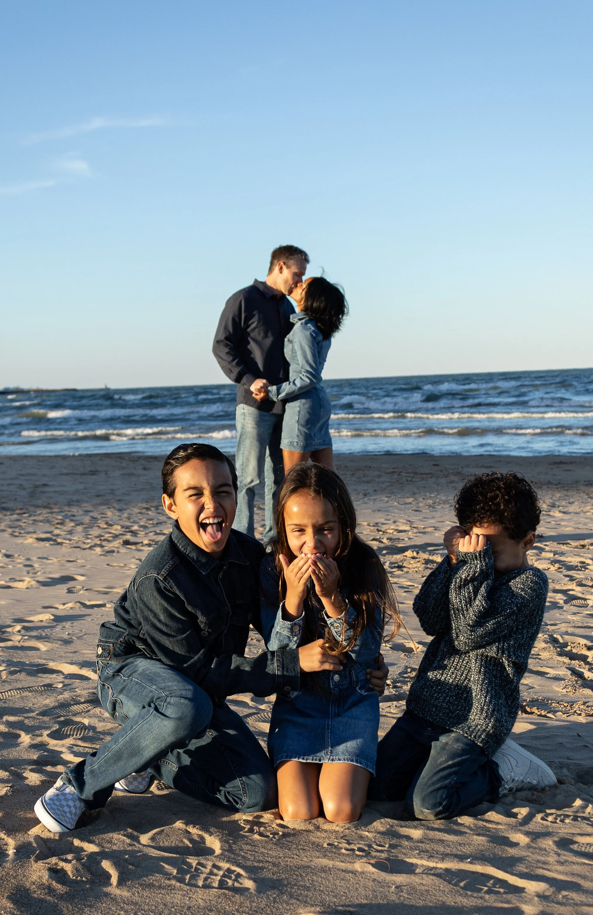 A group of children and teenagers on the beach, some sitting and some standing, enjoying a sunny day by the ocean with a clear blue sky.