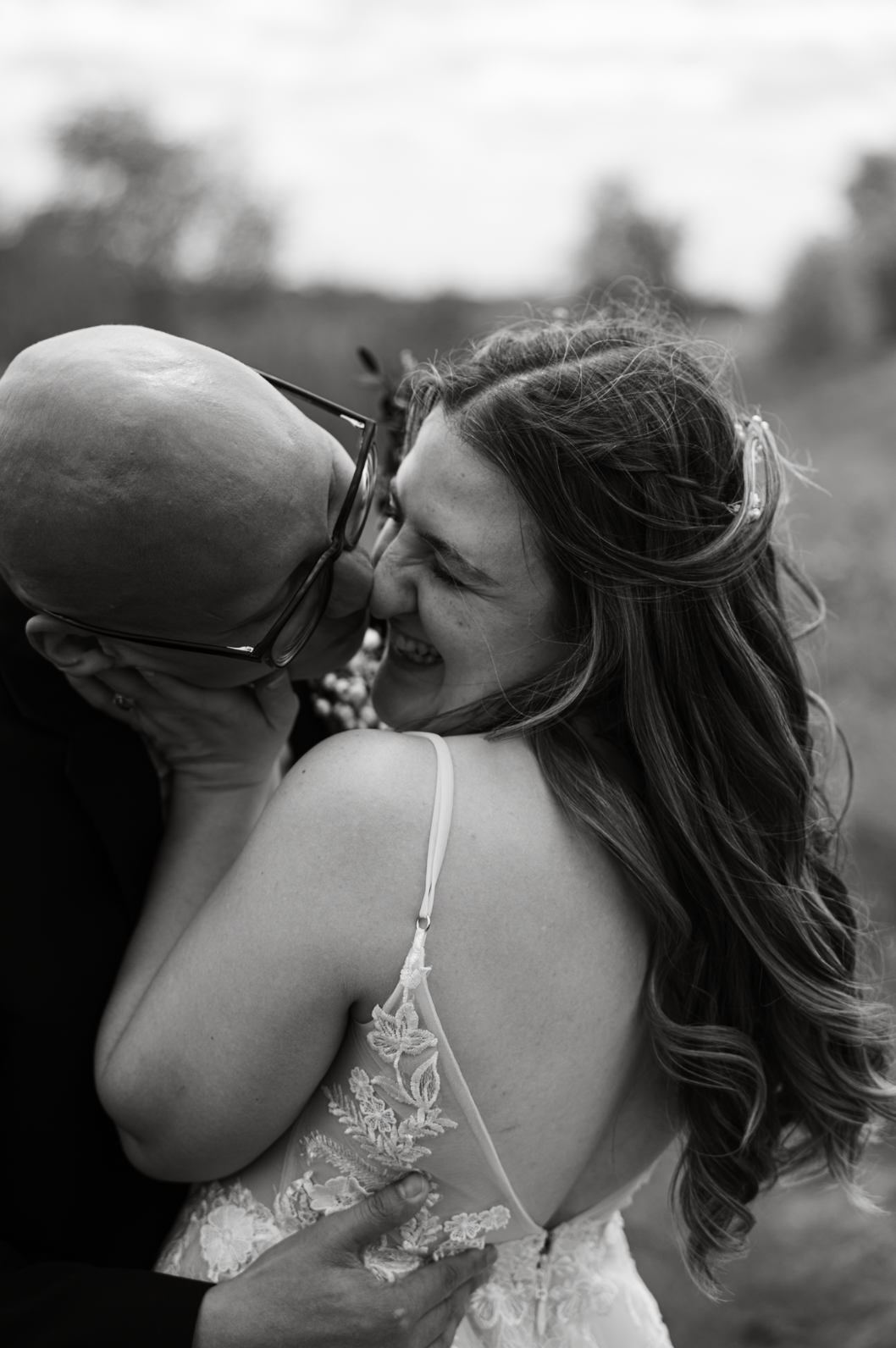 A black and white photo of a couple sharing a joyful moment, about to kiss outdoors. The woman has long, wavy hair and is wearing a spaghetti strap dress with floral embroidery. The man is bald, wearing glasses, and holding her close.