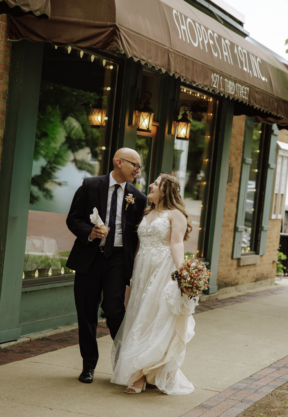 A couple in wedding attire walking outside a storefront, with the groom holding an ice cream cone, both smiling at each other.