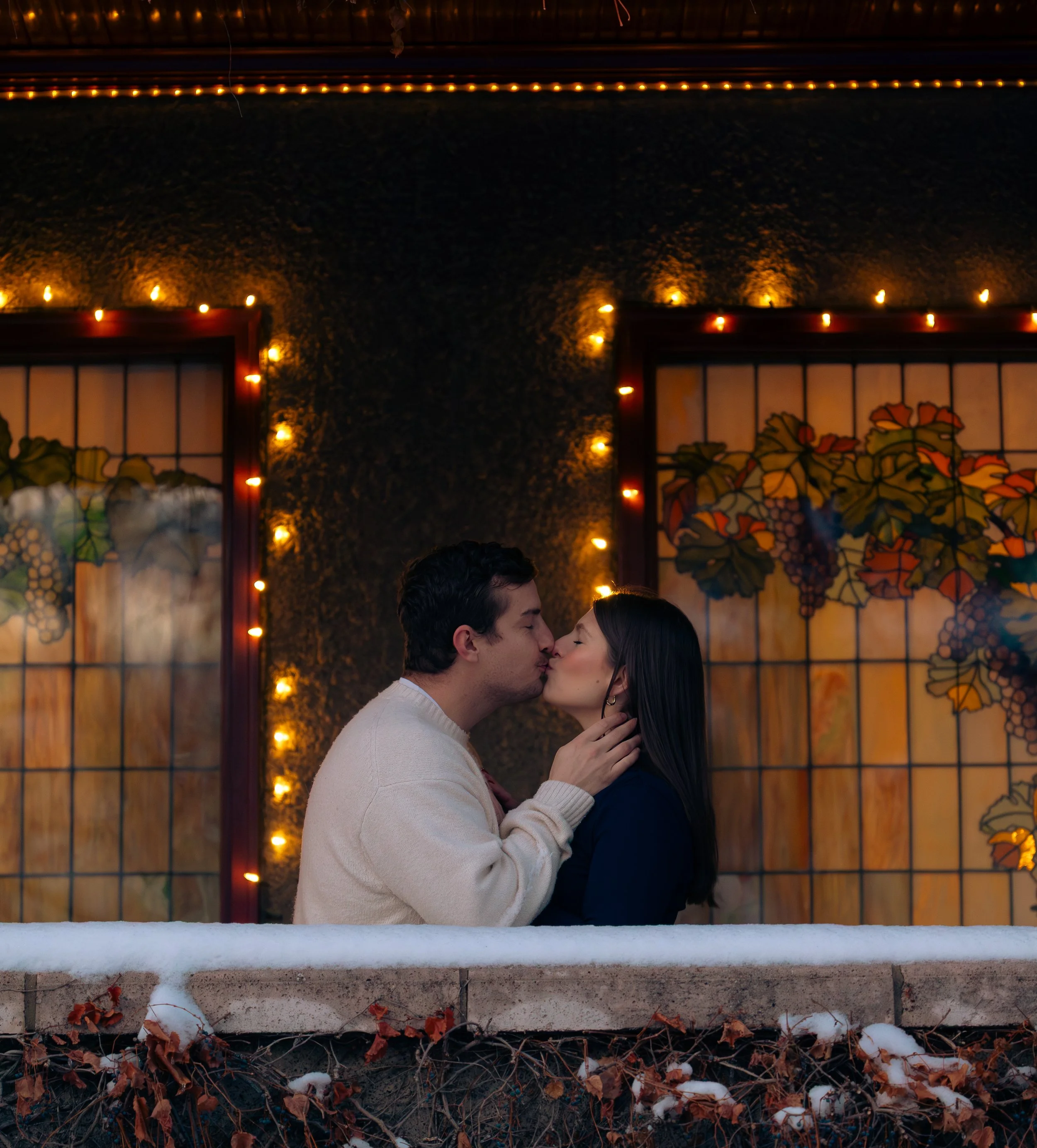 A couple is kissing in front of a stained glass window with a grapevine design, illuminated by string lights, outdoors during fall with snow and fallen leaves below.