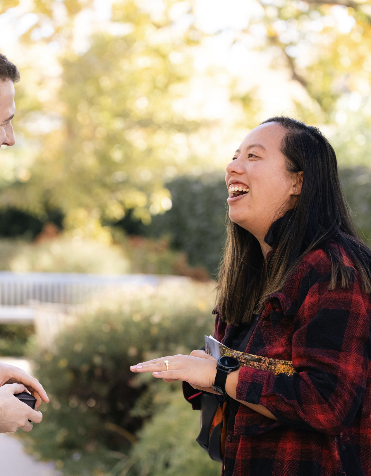 A woman with dark hair, wearing a black shirt and red plaid jacket, smiling and talking to a man outdoors during daytime, with trees and a bench in the background.