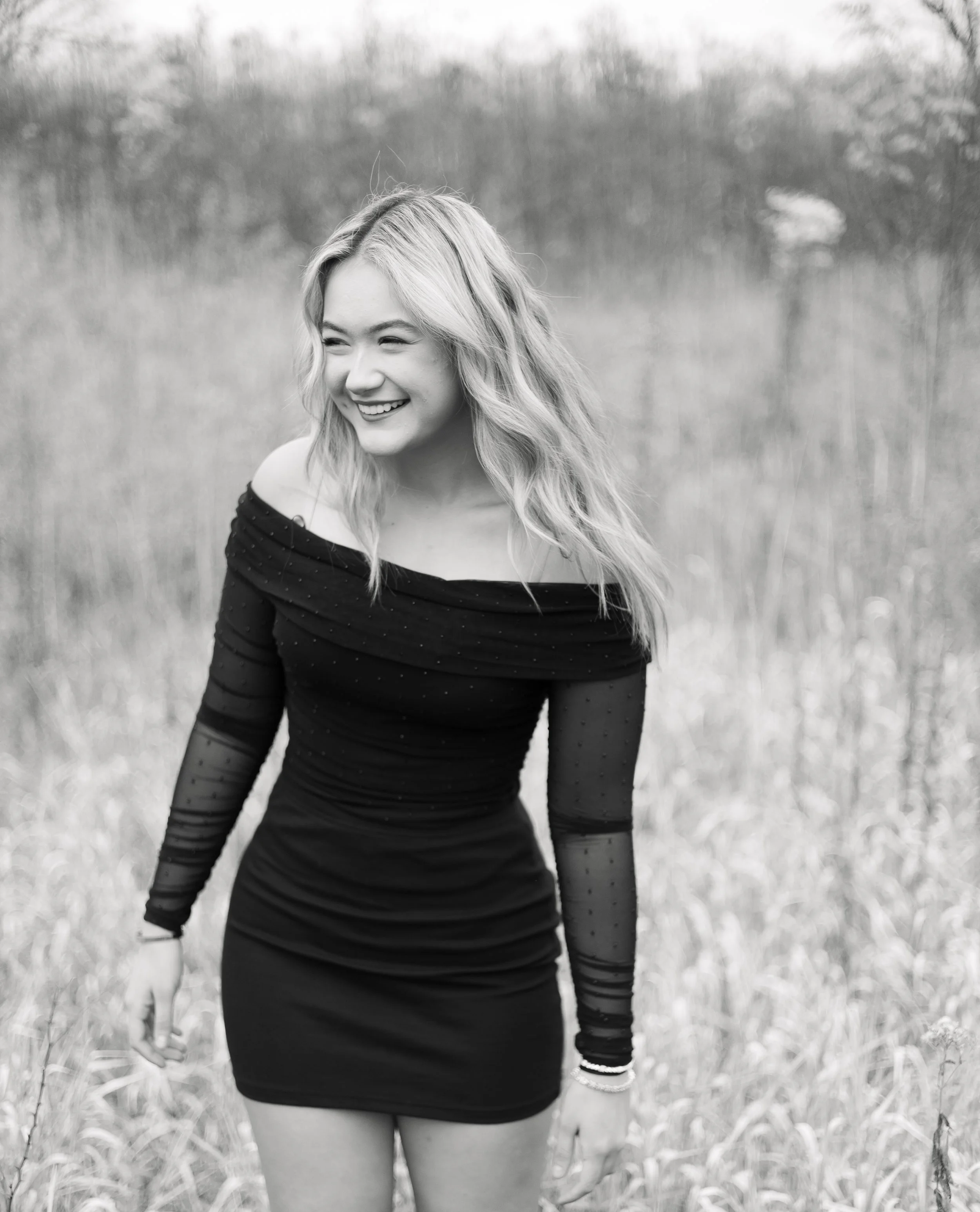 A black-and-white photo of a woman with long, wavy hair smiling and looking to the side, standing in a field with tall grass and trees in the background.