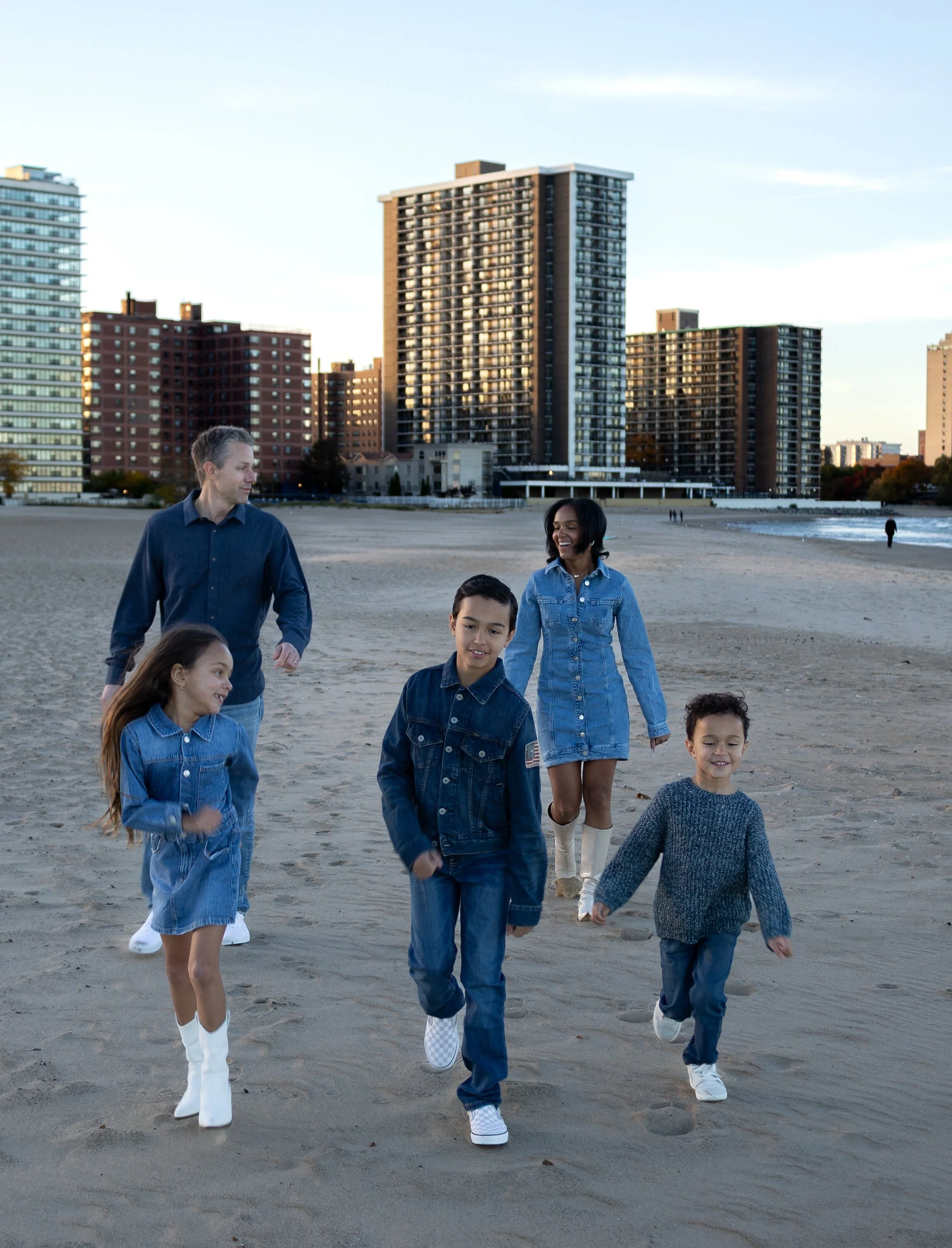 A group of diverse people walking on a sandy beach with tall apartment buildings in the background during sunset.