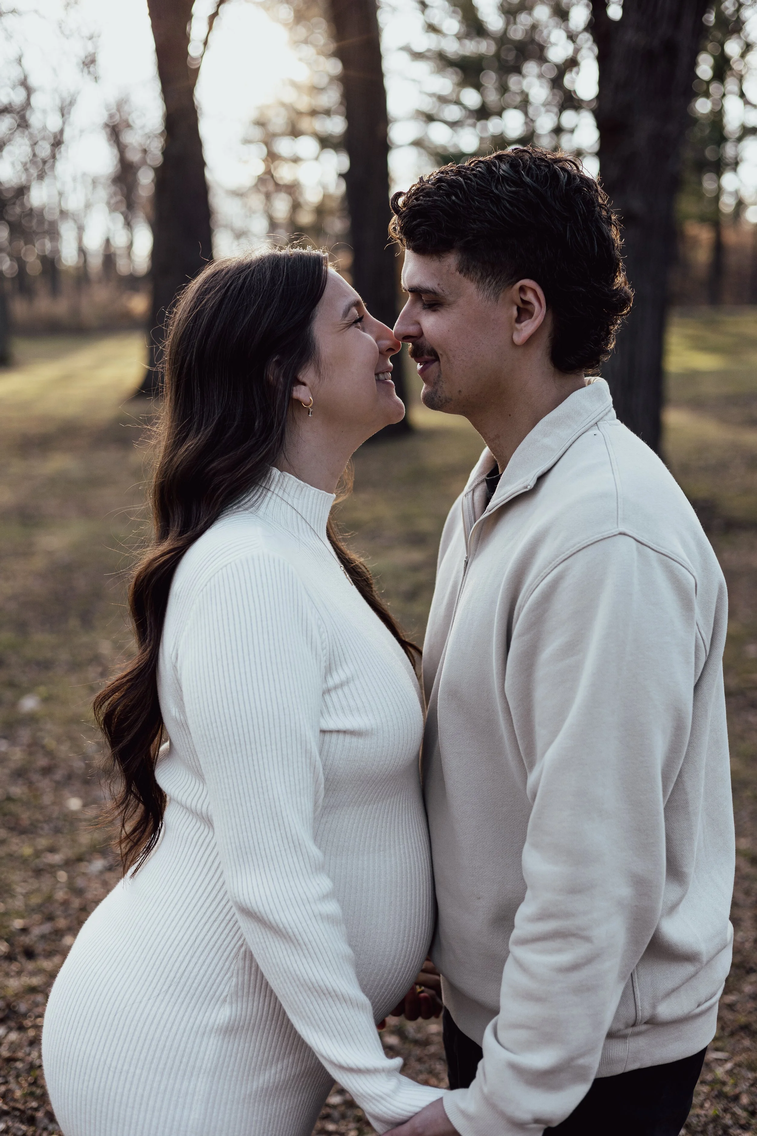 A couple stands close, smiling and touching noses in a wooded outdoor setting during sunset.
