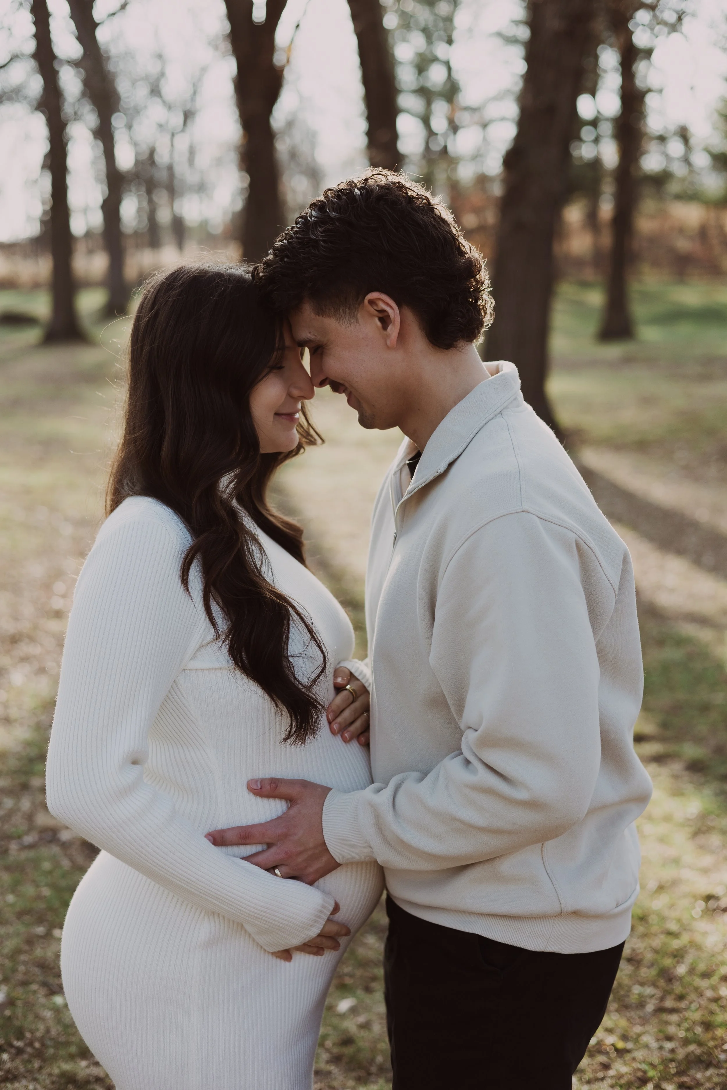 A couple standing outside in a park with trees in the background, pregnant woman and man touching foreheads, smiling, woman wearing a white ribbed dress, man in a white hoodie.