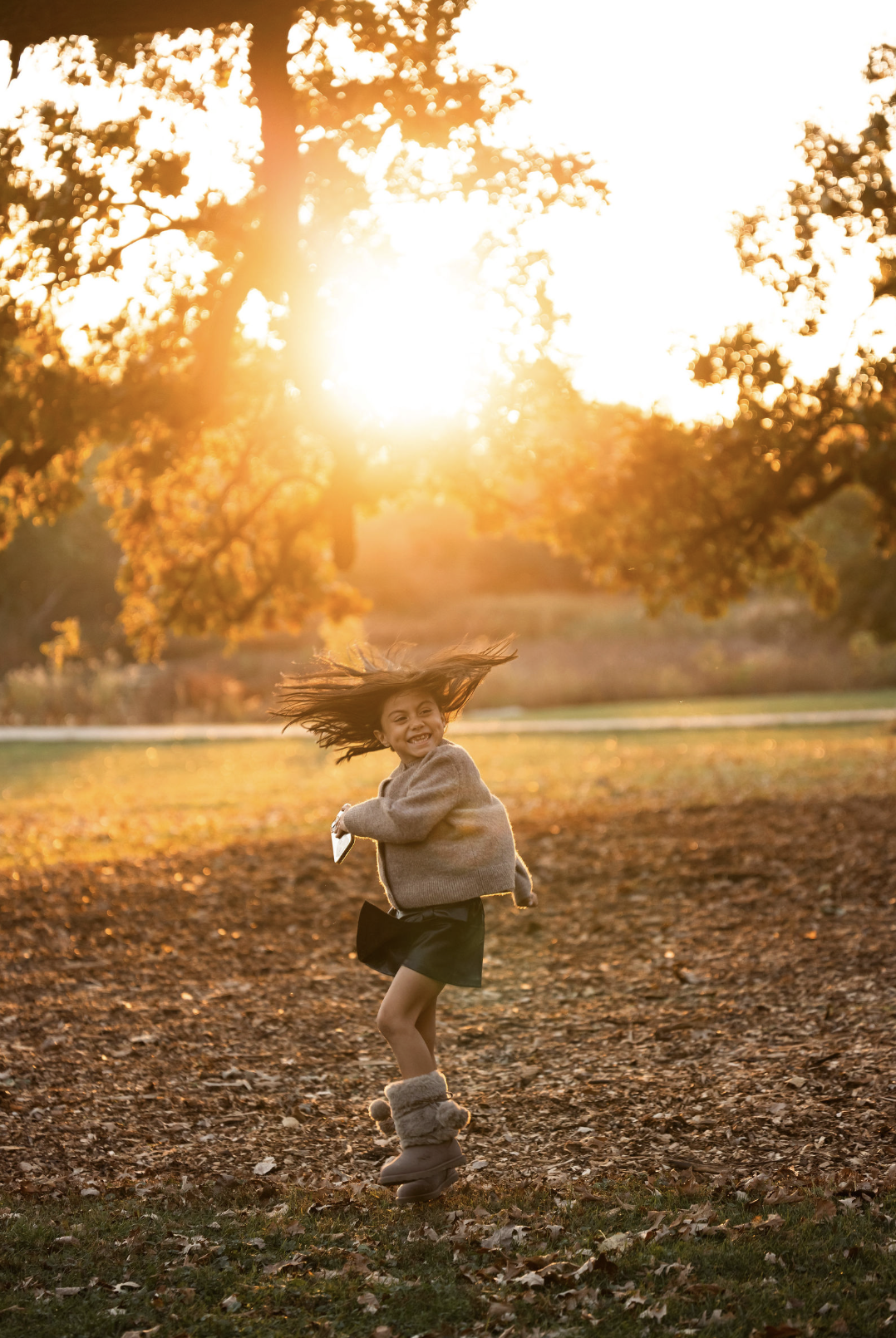 A young girl joyfully spinning on a park trail during golden hour, with falling leaves and trees in the background.