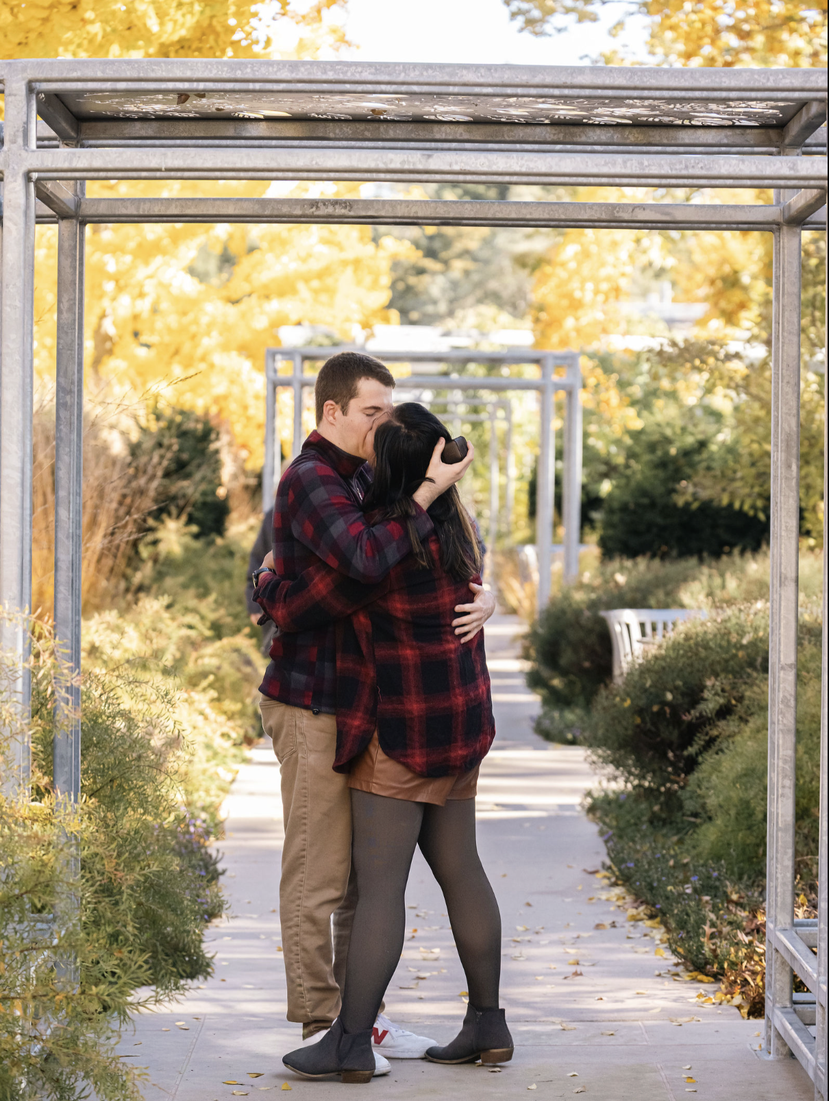 A young couple hugging and sharing a kiss under a metal archway on a sidewalk in autumn, with yellow and orange trees in the background.