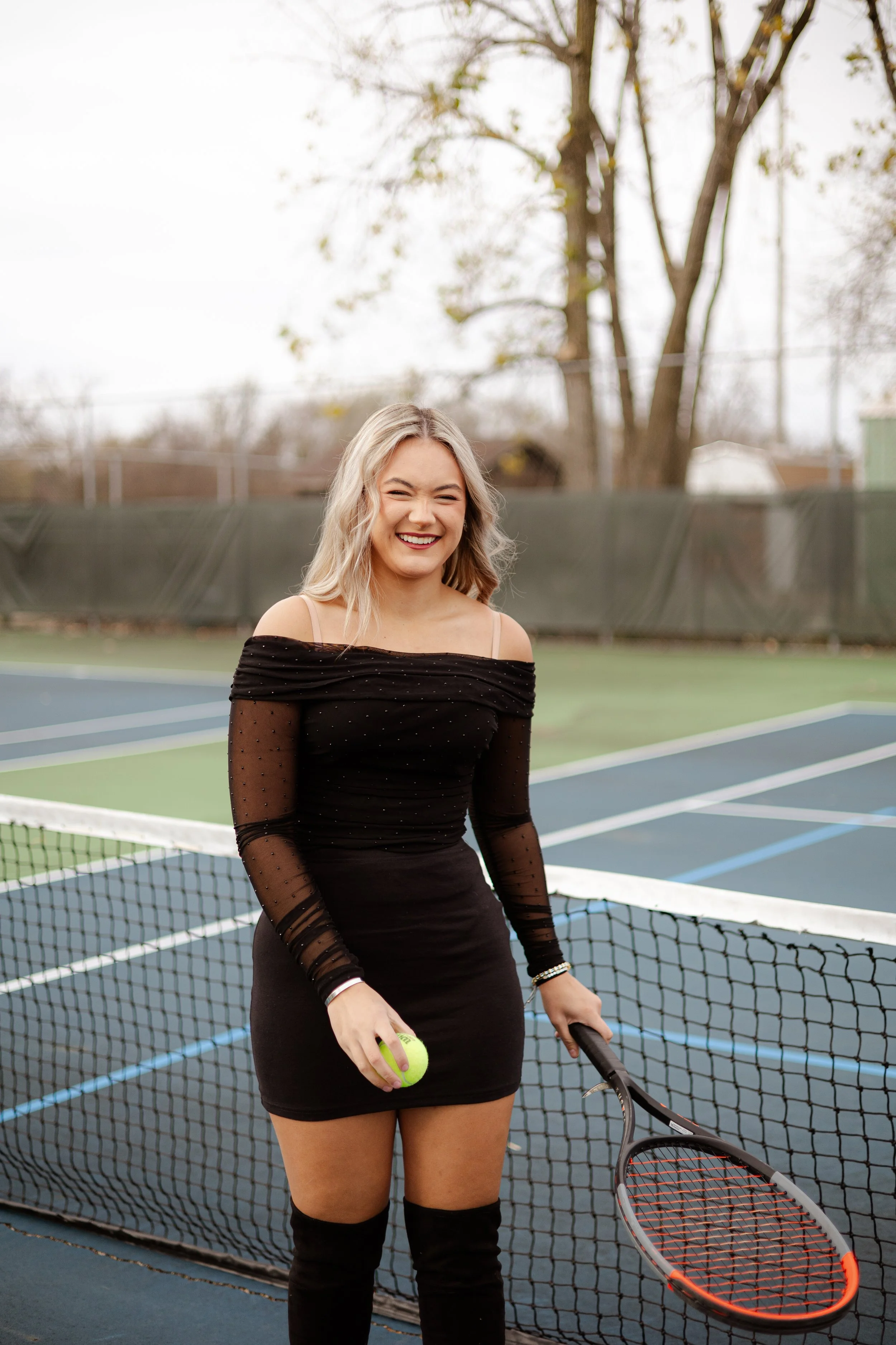 Young woman with blonde hair, wearing a black off-shoulder dress with sheer, dotted long sleeves, standing on a tennis court holding a tennis ball and tennis racket, smiling and squinting, with a net in front and trees in the background.