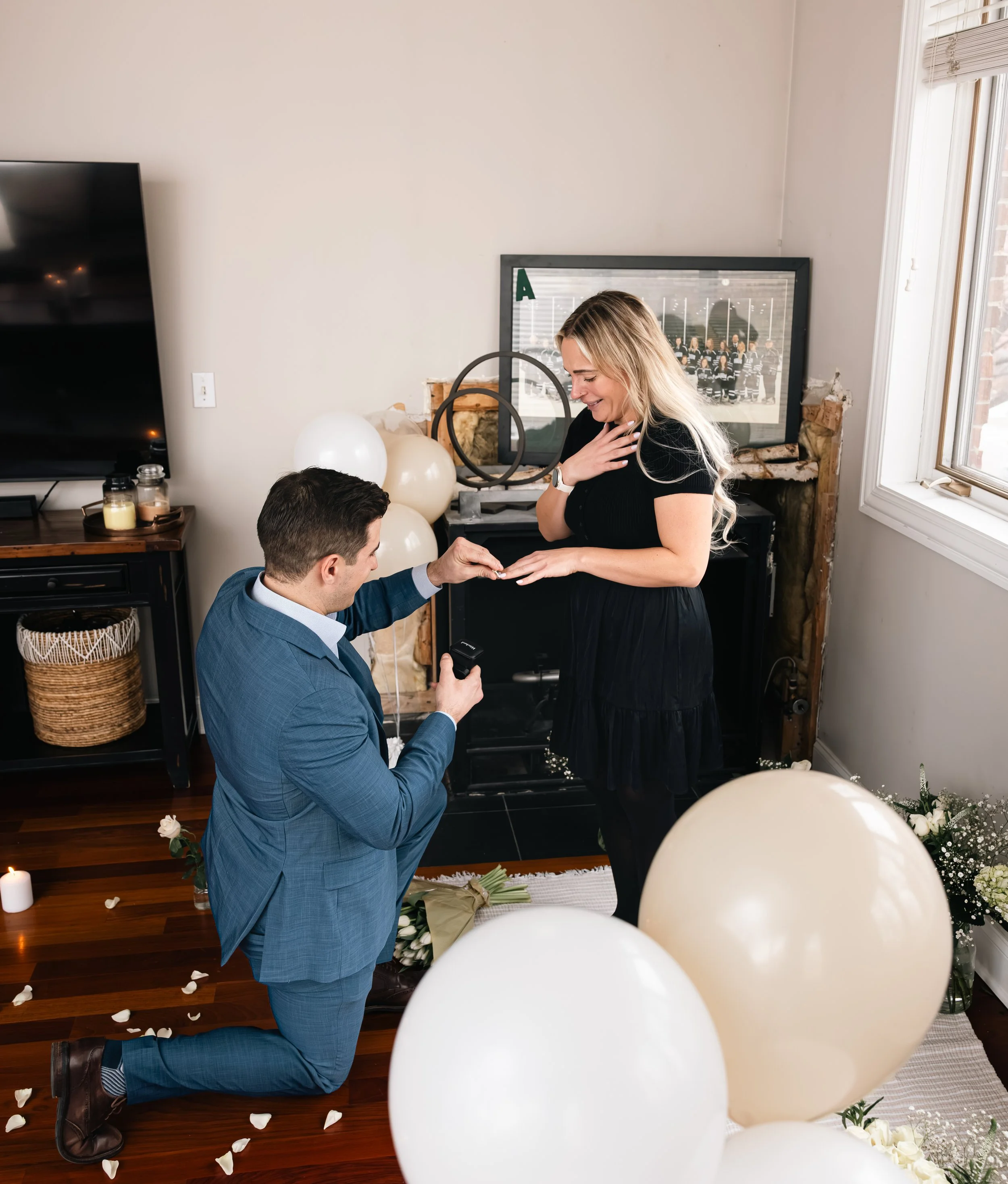 A man kneeling and proposing marriage to a woman in a decorated room with white balloons, flower petals, candles, and a wedding photo in the background.