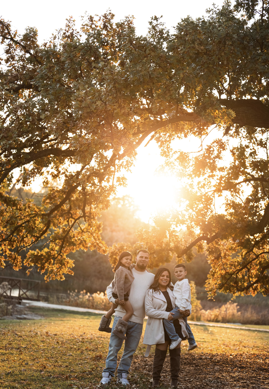 A family of four standing outdoors during fall at sunset. Two parents and two children are smiling, with trees with autumn leaves and a pond in the background.
