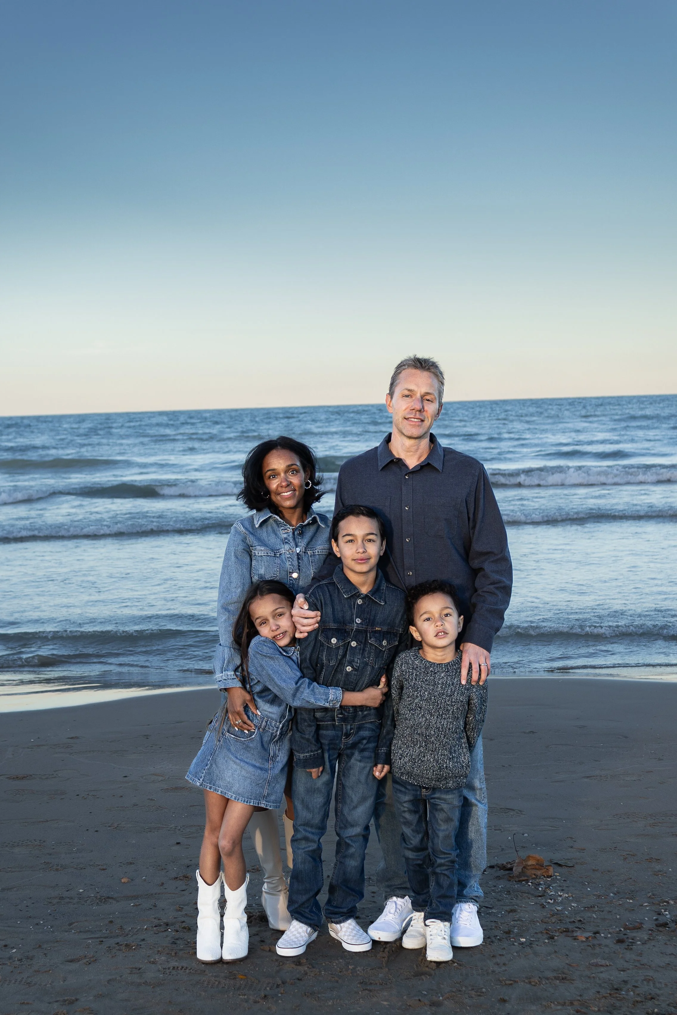 A family of seven, with three children and four adults, standing on a beach near the ocean as the sun sets or rises in the background, all dressed in casual denim and comfortable clothing.
