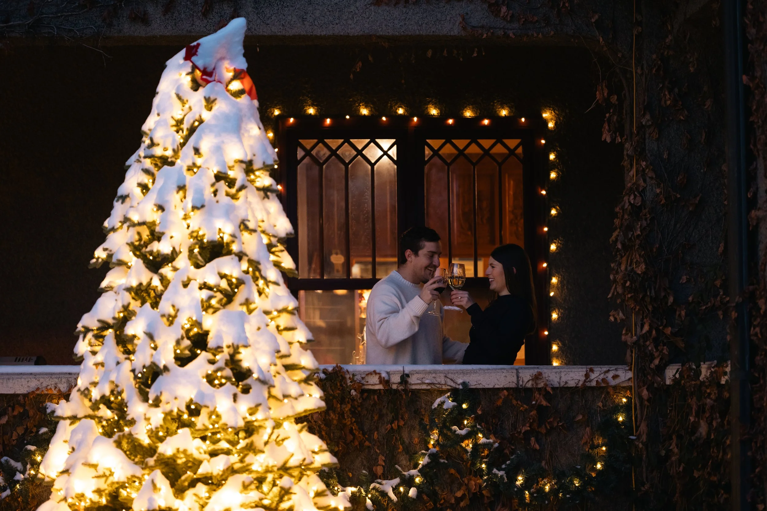 A couple toasting with glasses of wine in front of a snow-covered decorated Christmas tree and a warmly lit window at night.